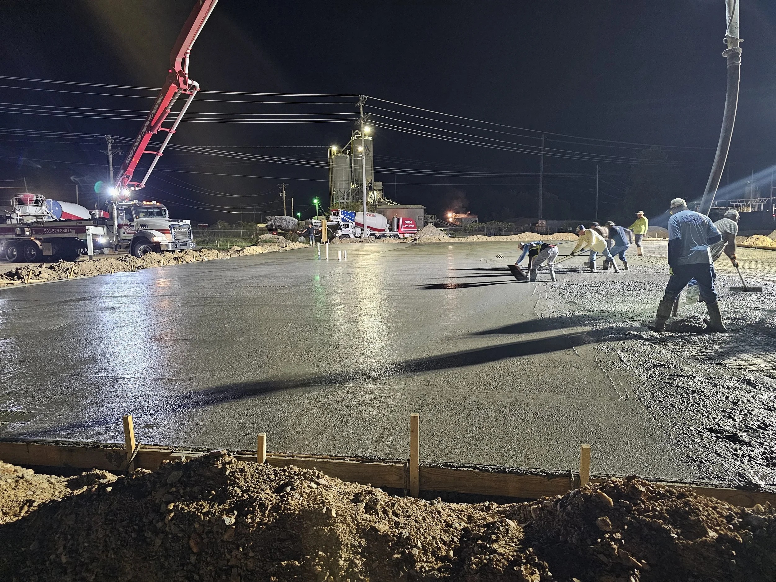 Nighttime construction site with workers pouring concrete, several cement mixers, a crane, and power lines visible in the background.