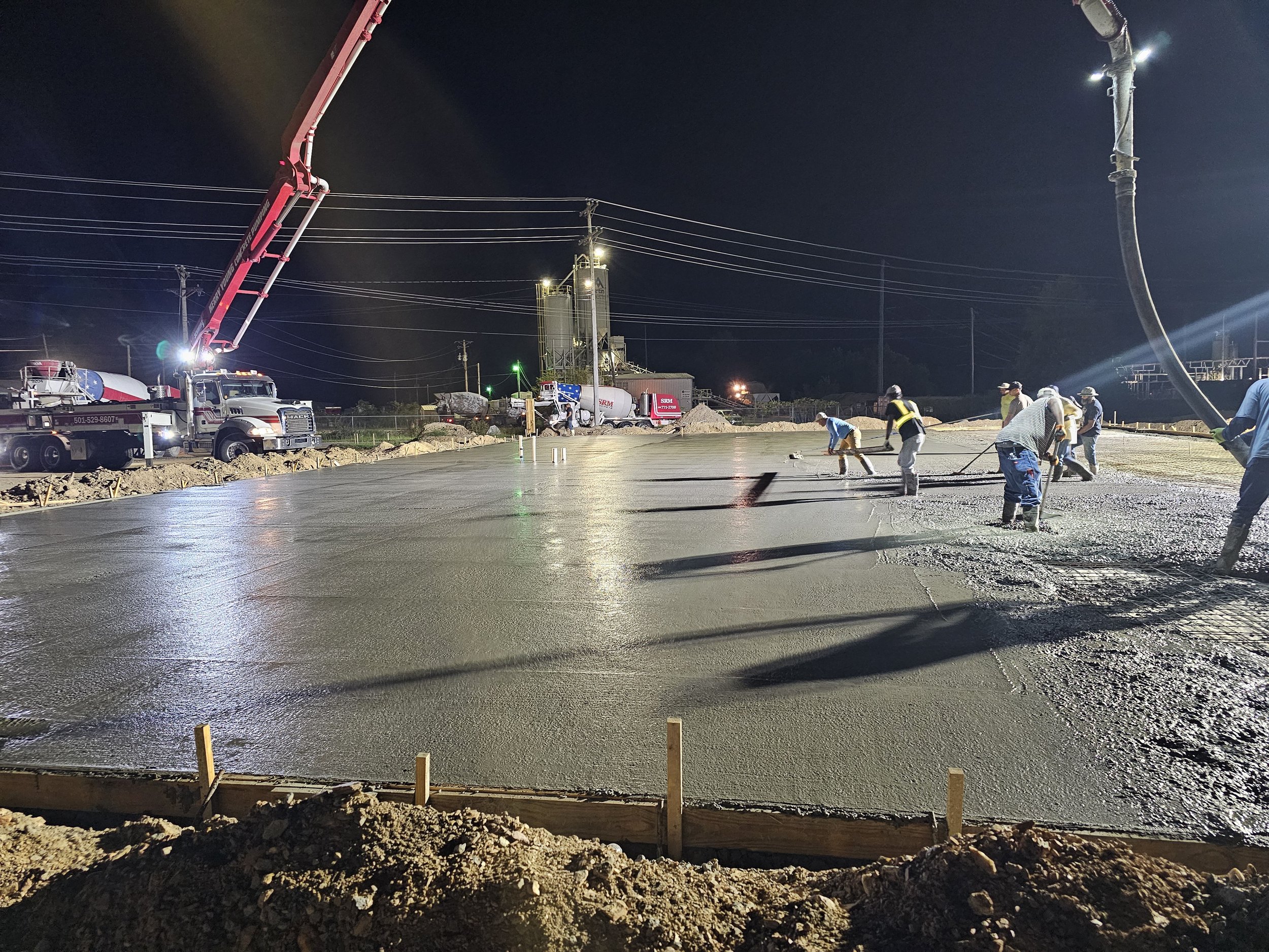 Workers are pouring concrete on a construction site at night, with machinery and utility poles in the background, and the scene is illuminated by artificial lighting.