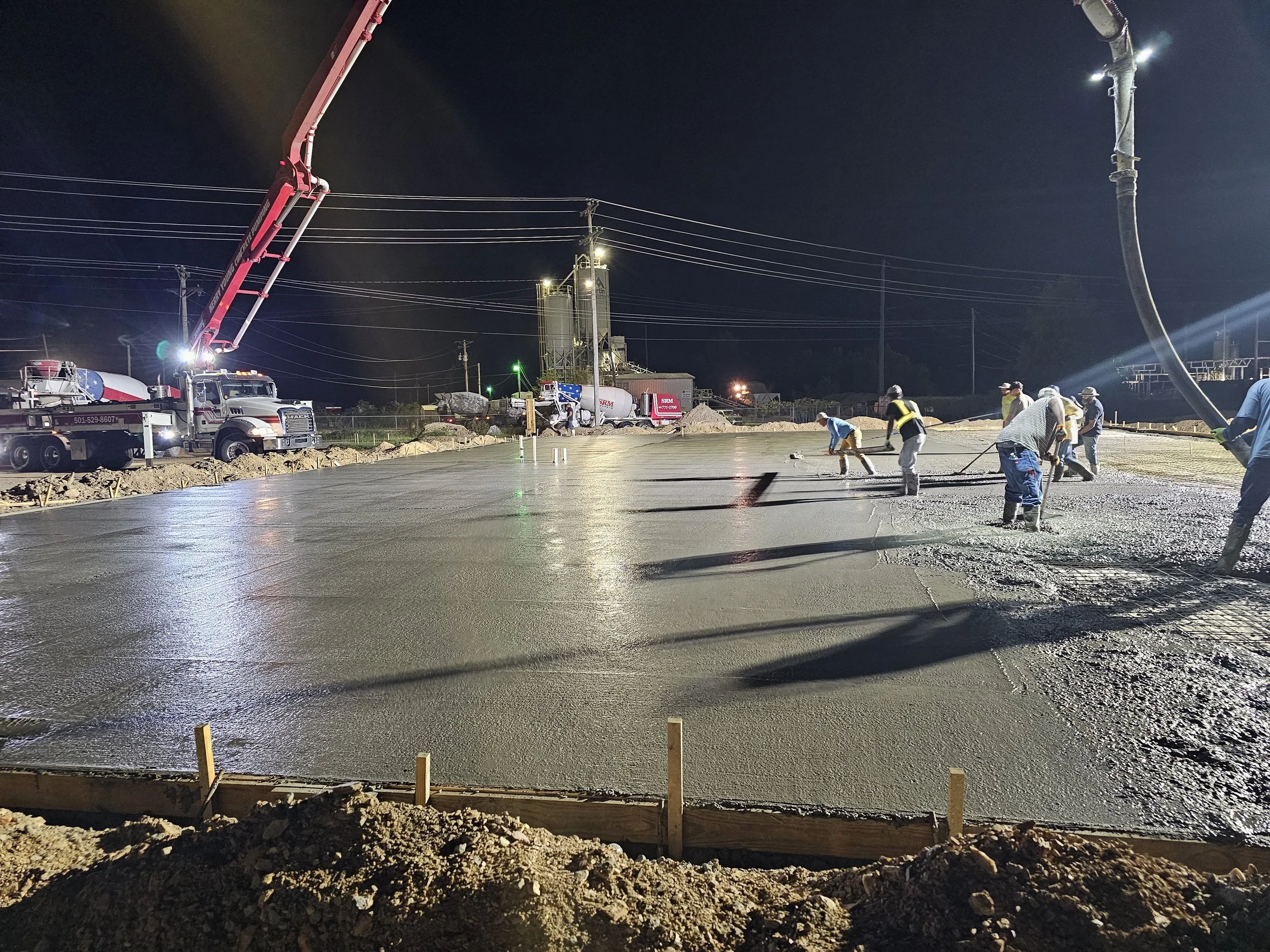 Workers are pouring concrete on a construction site at night, with machinery and utility poles in the background, and the scene is illuminated by artificial lighting.