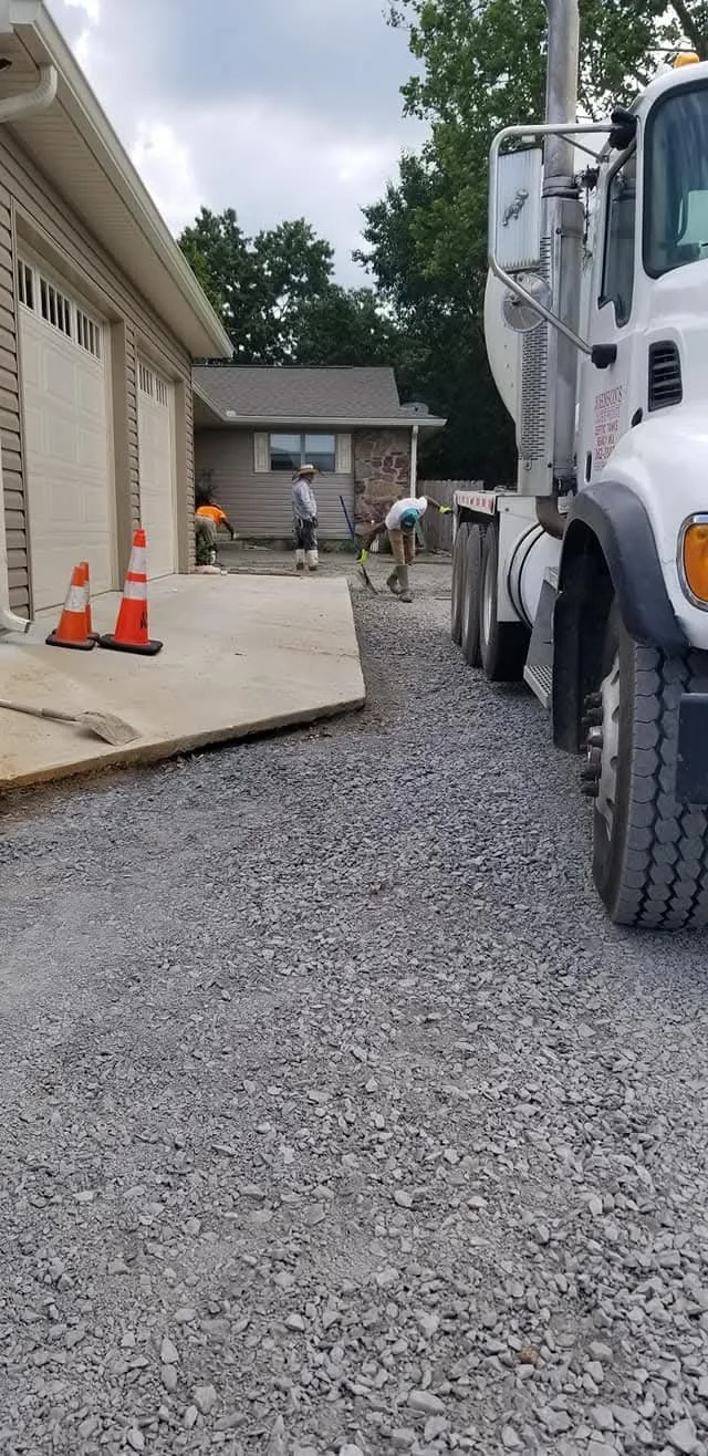 Construction workers leveling the ground and working on a pathway in front of a house, with a large truck parked nearby and traffic cones marking the work area.