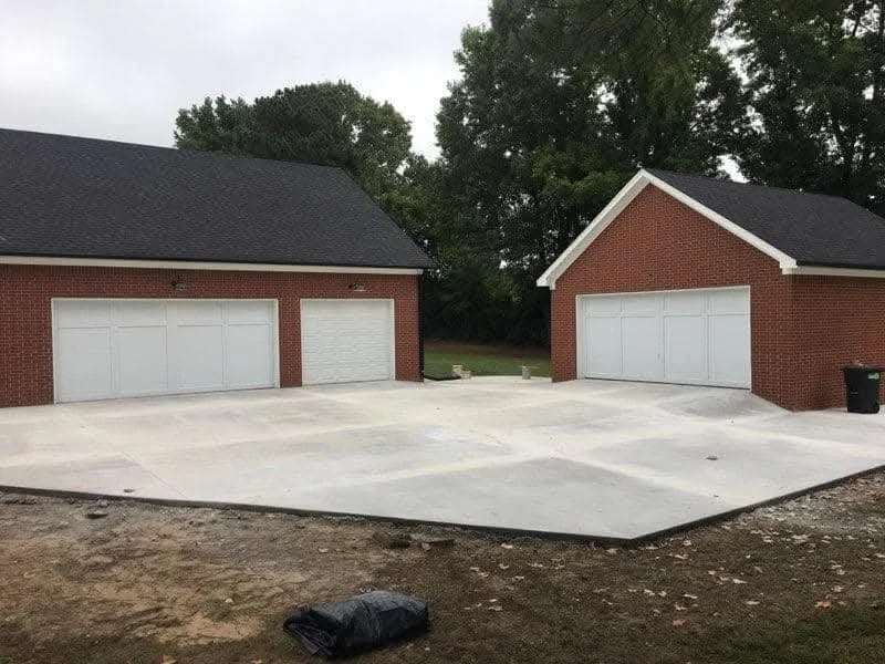 Two attached brick garages with white doors, one with an additional side door, and freshly poured concrete driveway in front.