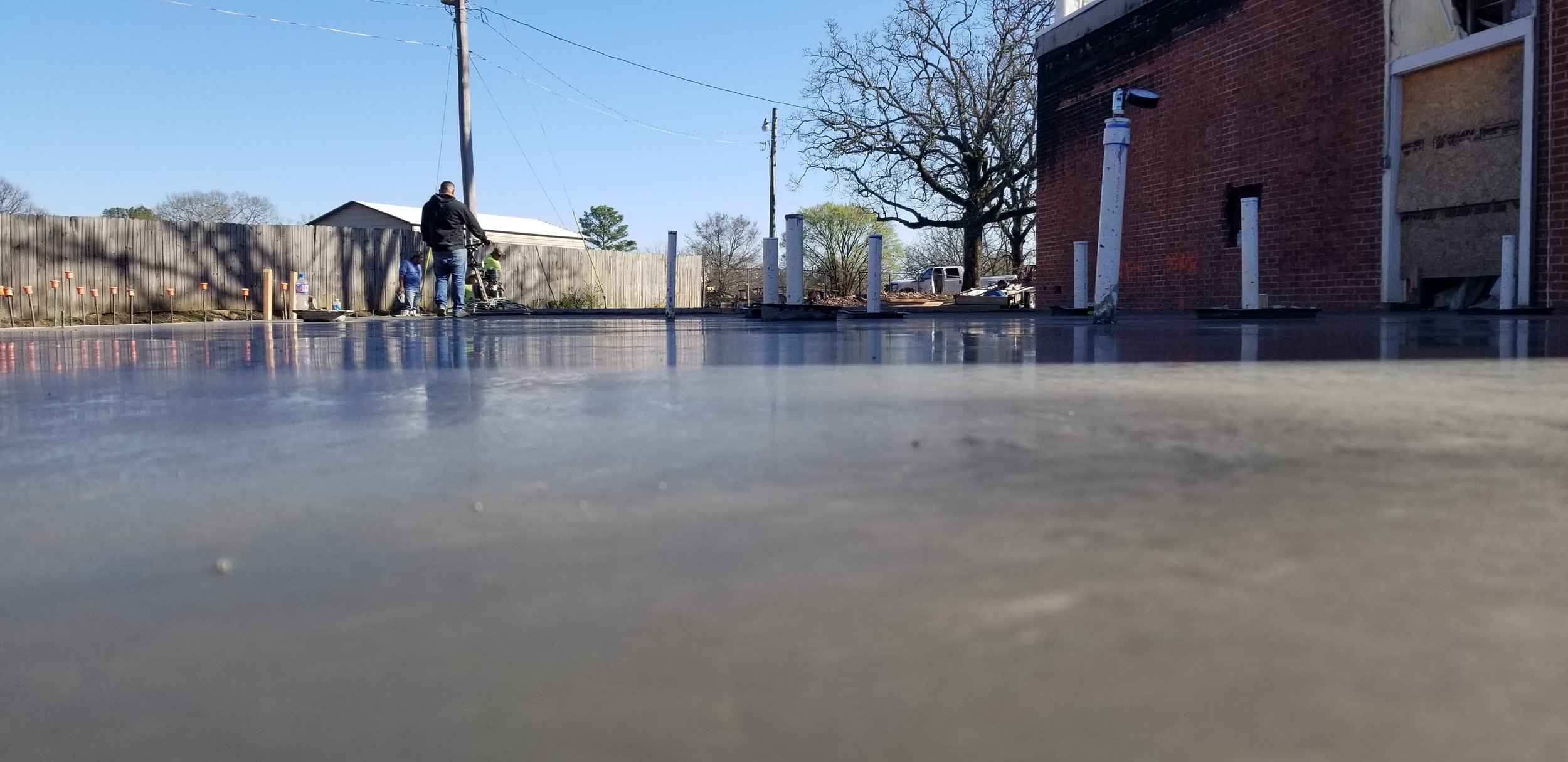 People working on a construction or renovation project outside, with a focus on a freshly poured concrete slab, a wooden fence, and a brick building, during a sunny day.