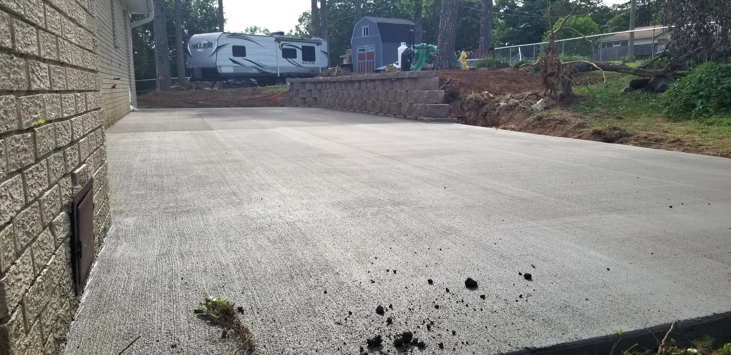 A newly poured concrete driveway next to a brick house wall, with some scattered dirt and small rocks in the foreground, and a backyard with trees, a small shed, a travel trailer, and a retaining wall in the background.
