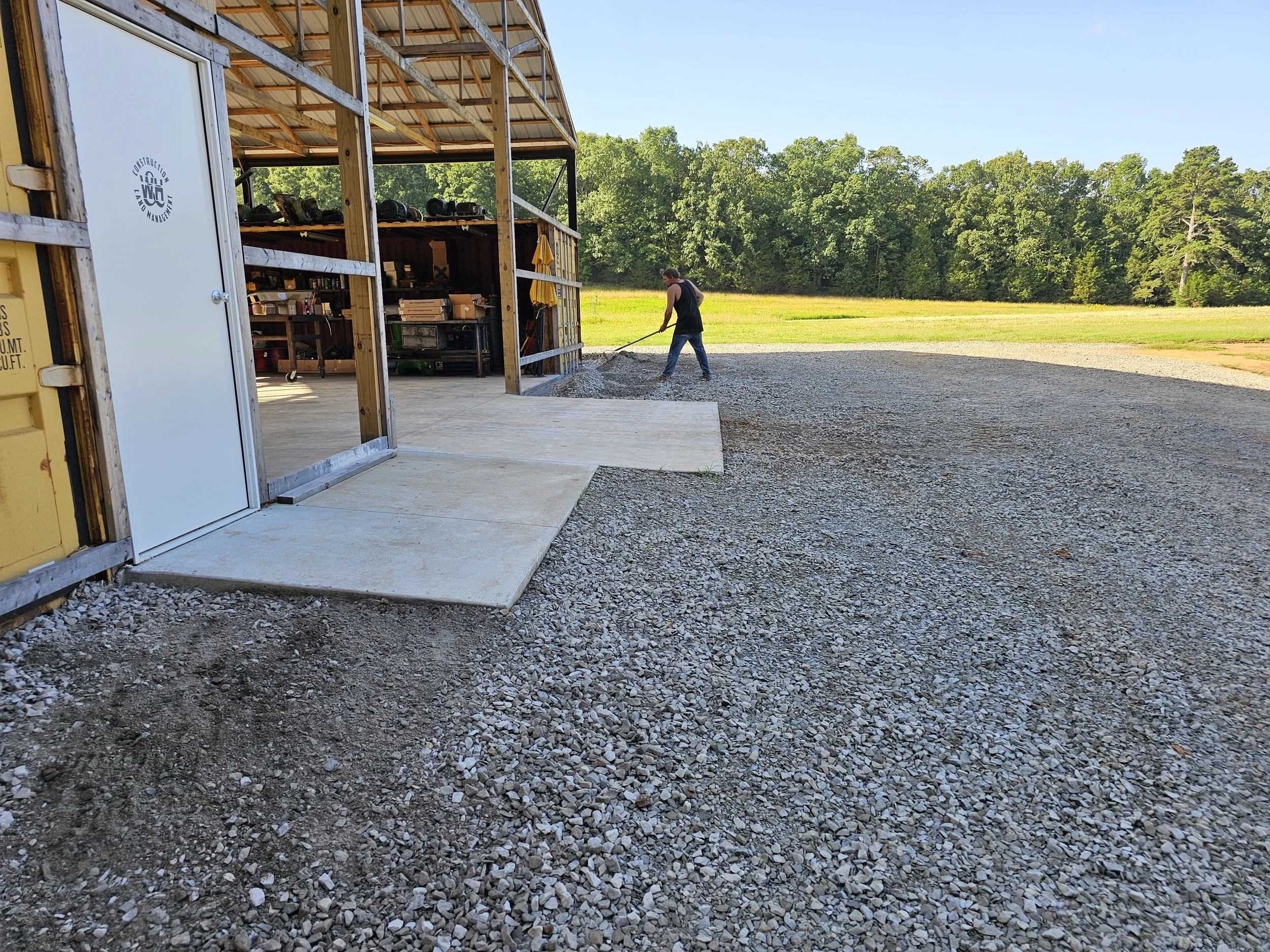 A man raking gravel outside a wooden structure with storage shelves, surrounded by greenery and trees under a clear blue sky.