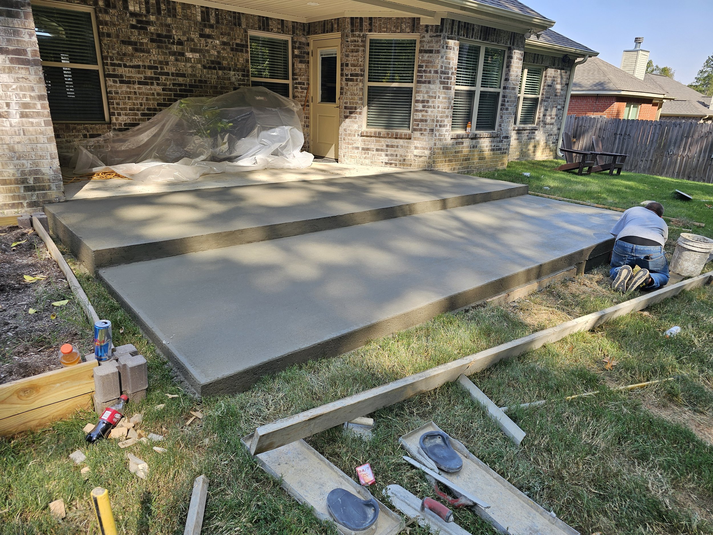 A person working on a concrete porch extension in a backyard during daytime. There are three levels of poured concrete, with construction tools and supplies scattered around. The house exterior is brick with multiple windows, and a wooden fence separ