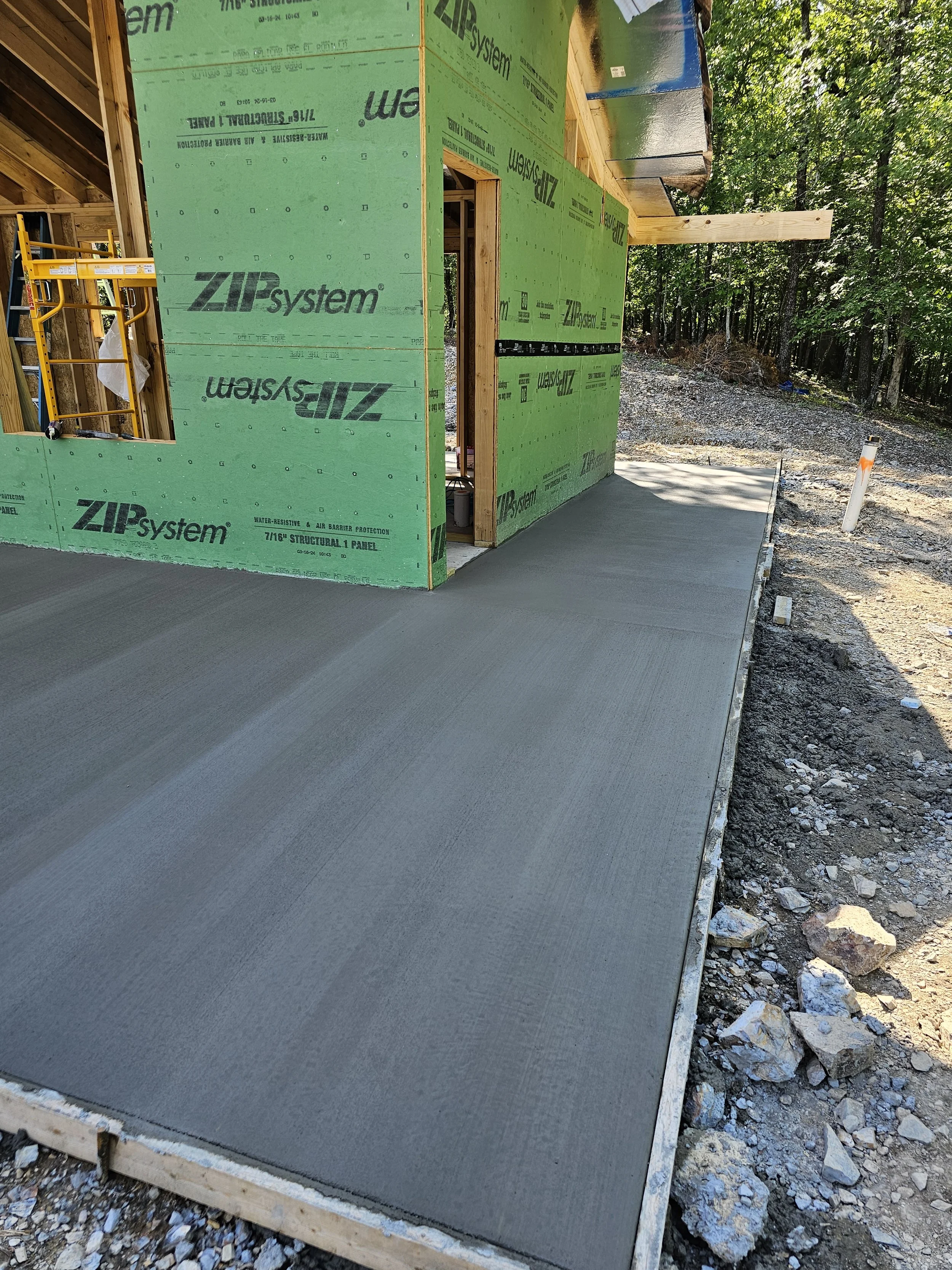 Construction site showing the foundation with a freshly poured concrete slab, wooden framing, and green weather-resistant panels on a building under construction in a wooded area.