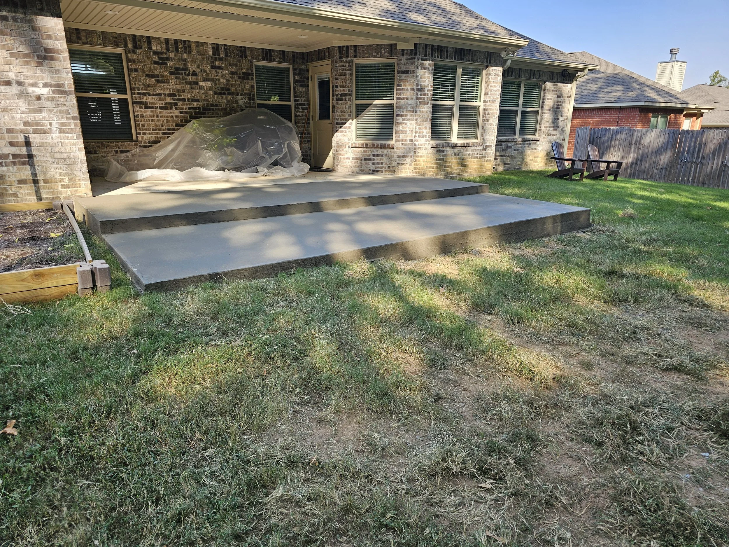 Backyard patio with concrete stairs, lawn, two wooden chairs, and brick house with large windows.