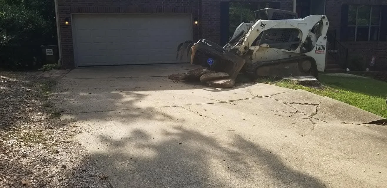 A small skid-steer loader parked in front of a residential garage, with a cracked driveway and some grass on the side.