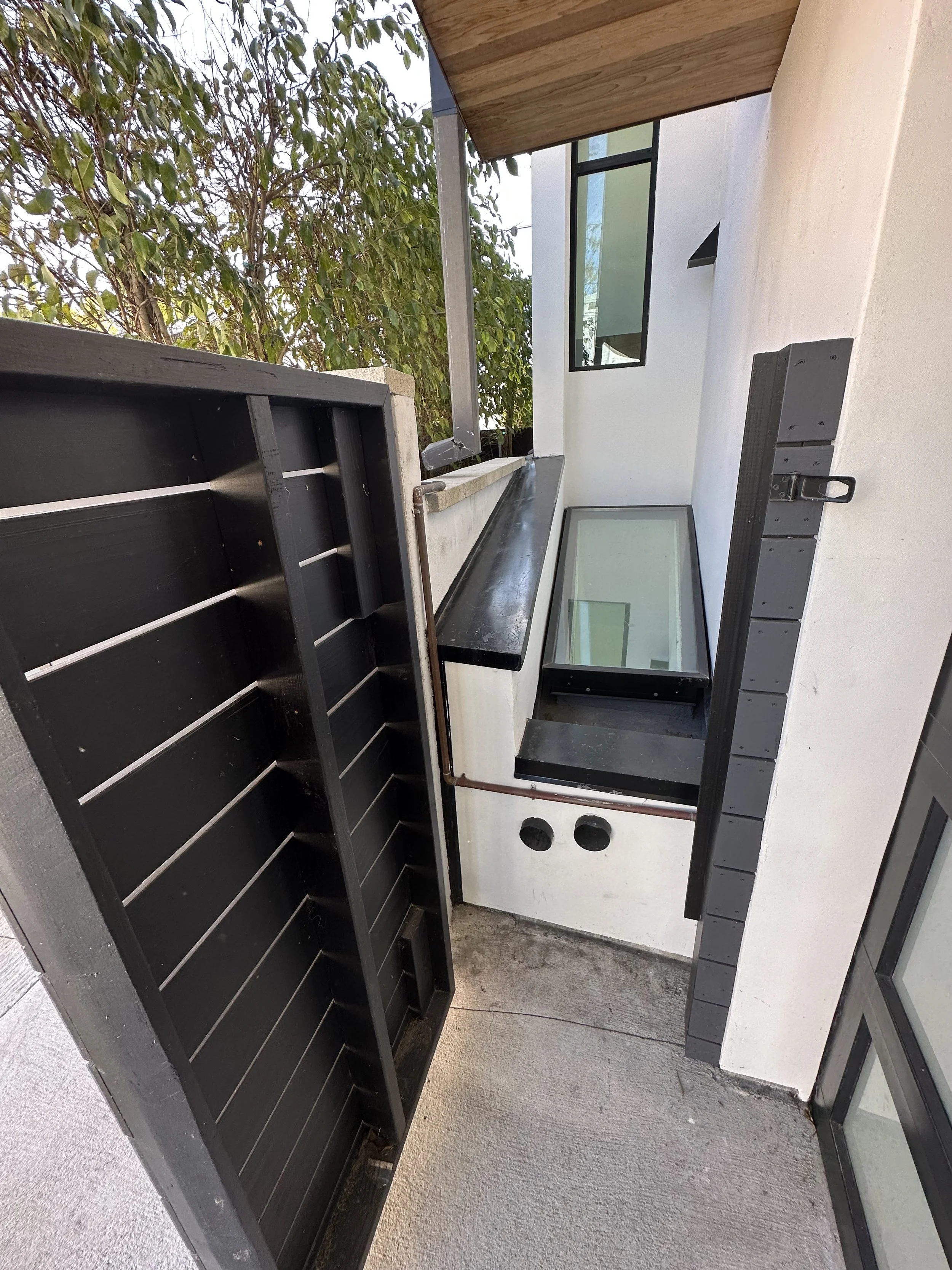 An outdoor balcony corner with a black railing, a glass window, a black countertop, and two round openings in the wall.