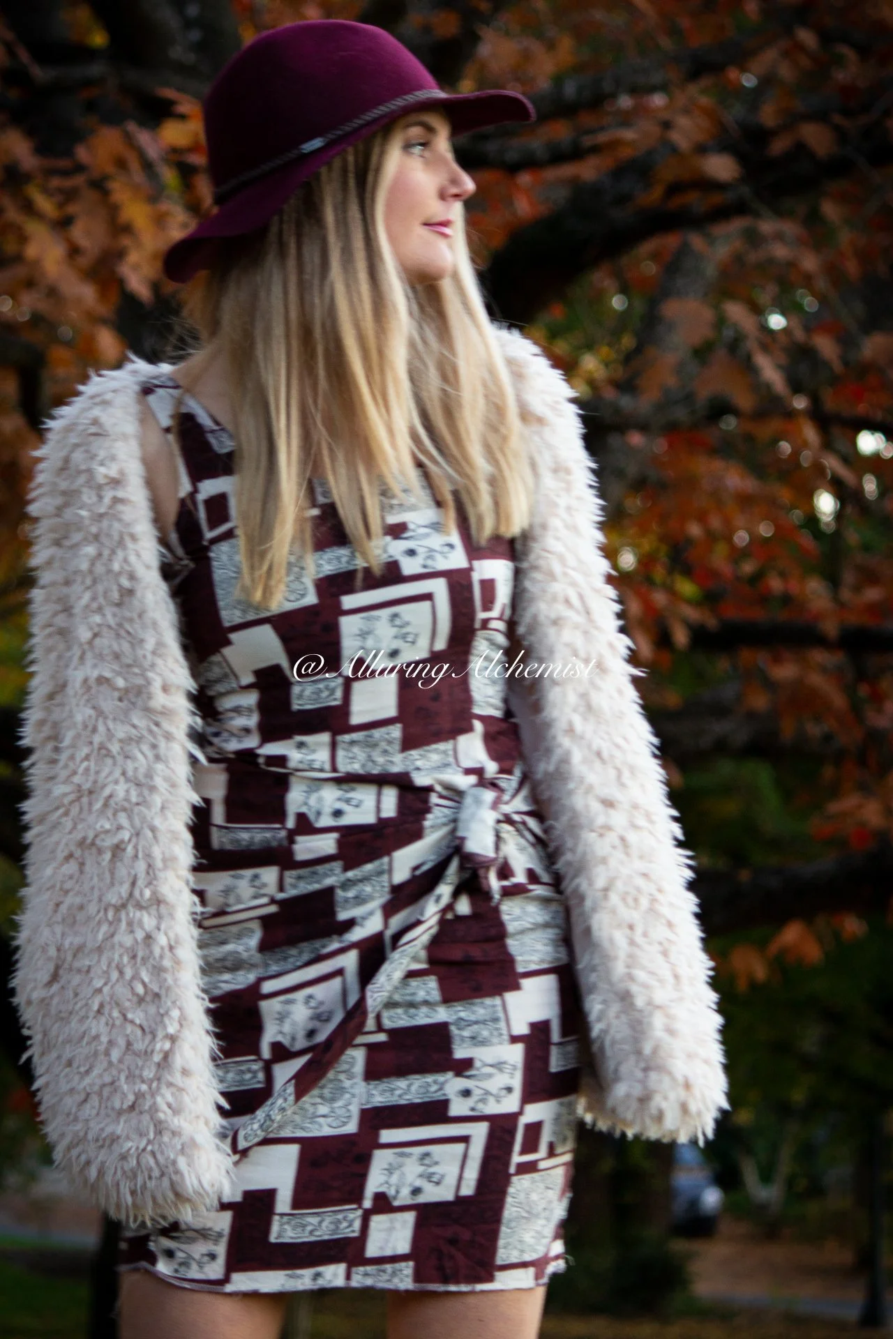 A woman wearing a maroon hat, patterned dress, and a fluffy beige shawl standing outdoors among autumn trees with orange leaves.