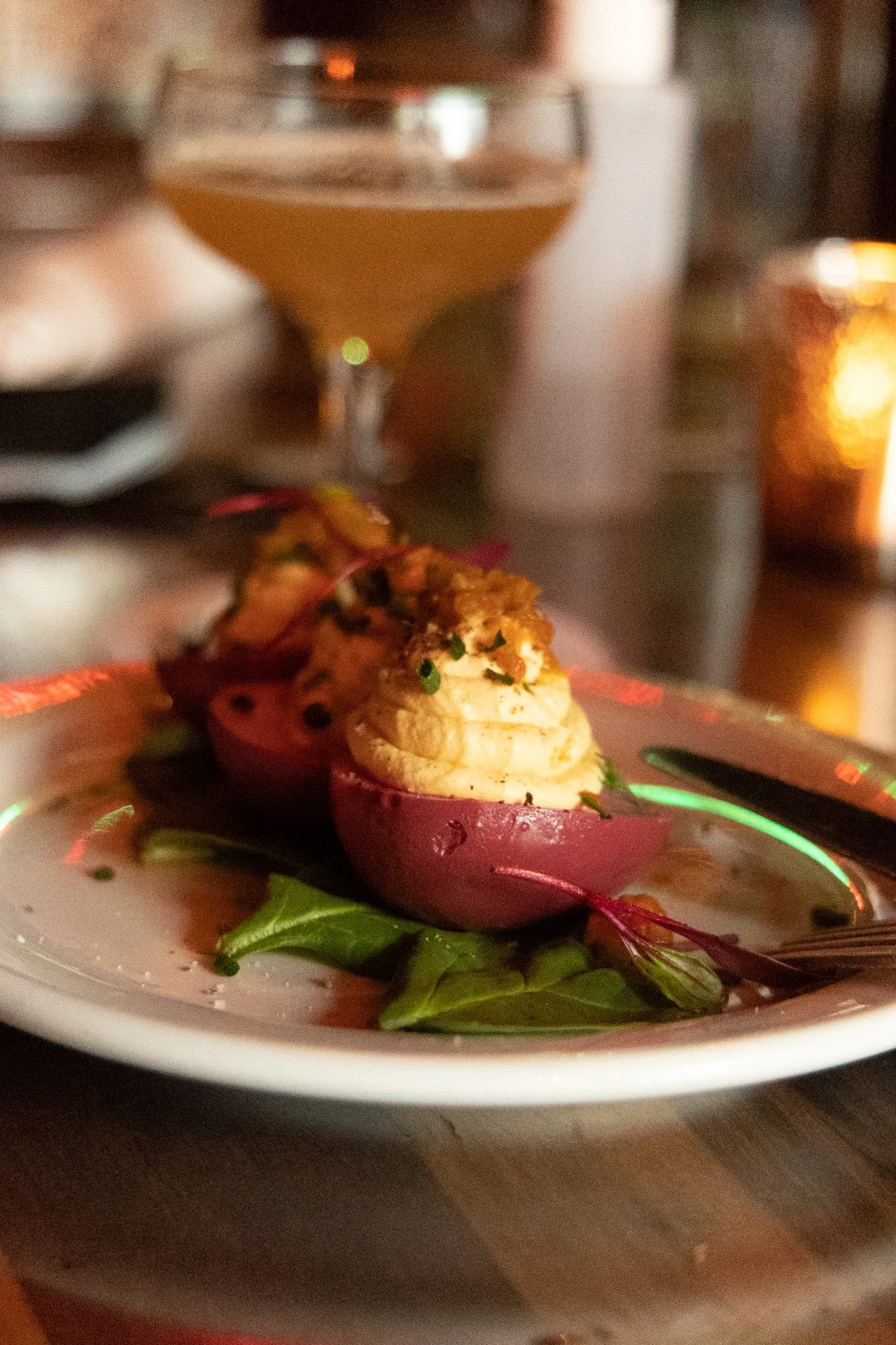 Close-up of a stuffed baked potato topped with herbs, served on a plate with greens, and a cocktail in a martini glass in the background.
