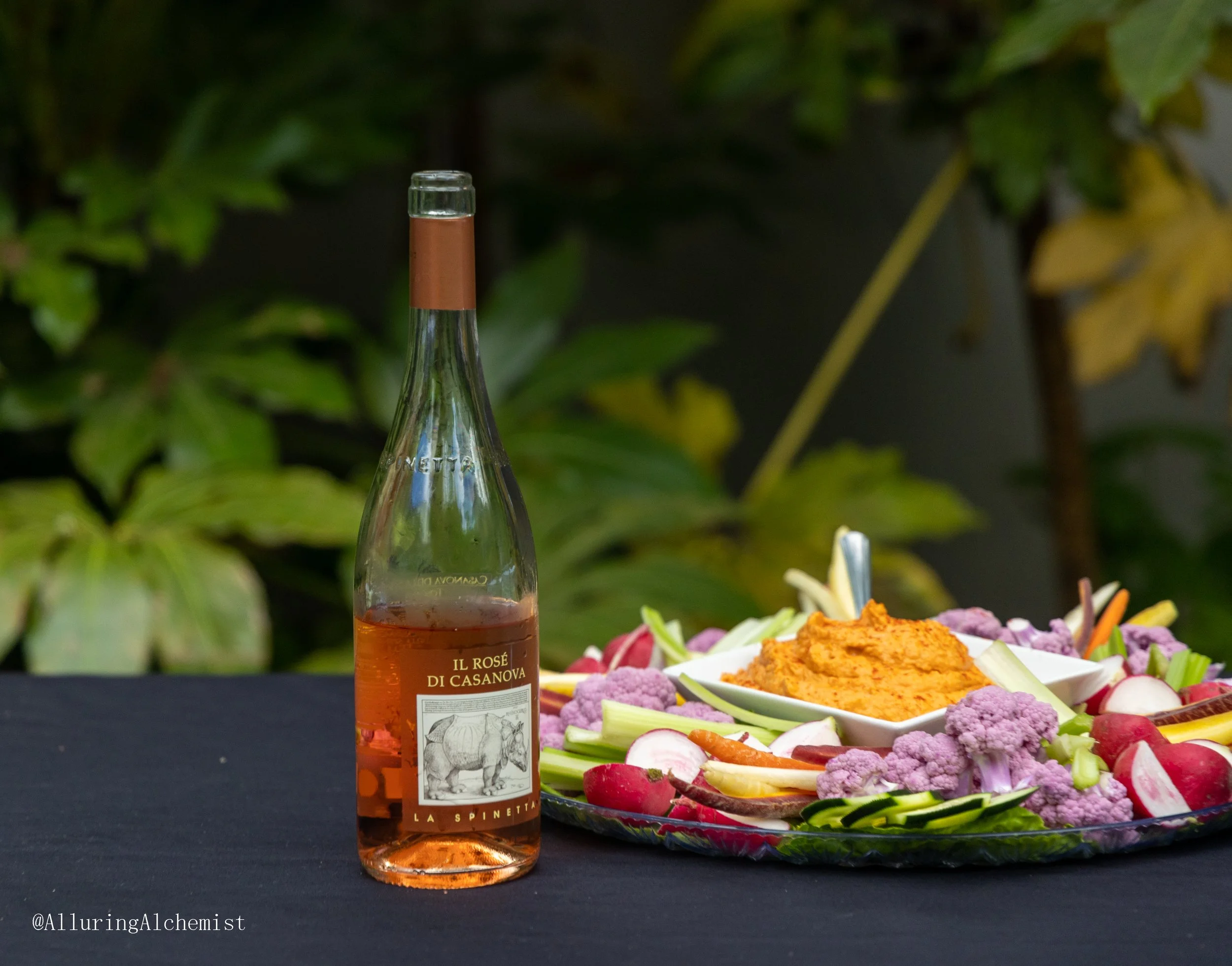 A partially visible bottle of Il Rosé di Casanova wine on a black table, surrounded by colorful vegetable and dip platter with radishes, cauliflower, and other vegetables, set outdoors with greenery in the background.