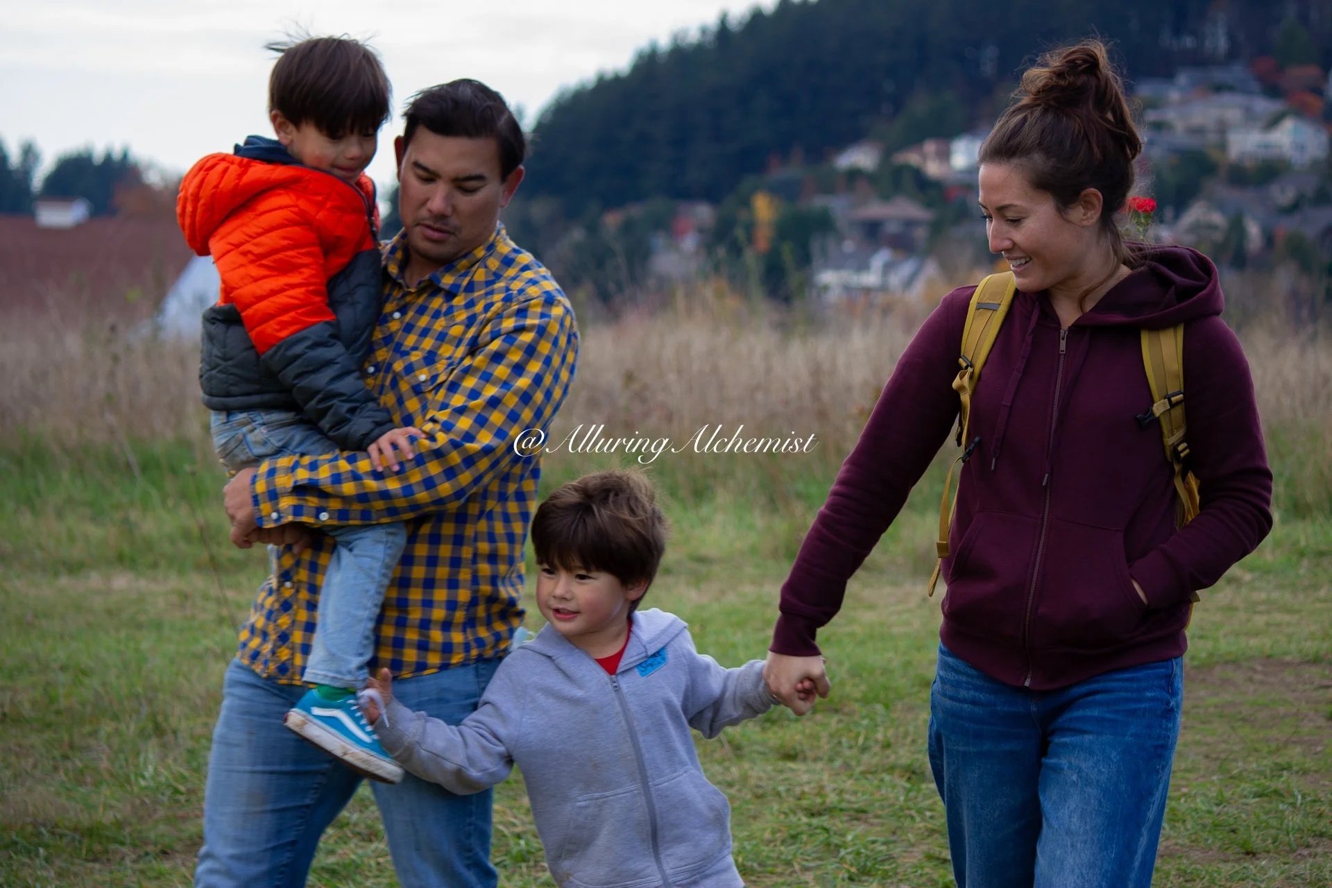 A family of three children and two adults walking outdoors in a grassy area with hills and houses in the background. One of the children is being held by a man in a yellow and blue checkered shirt, and another child is holding hands with a woman in a maroon hoodie.