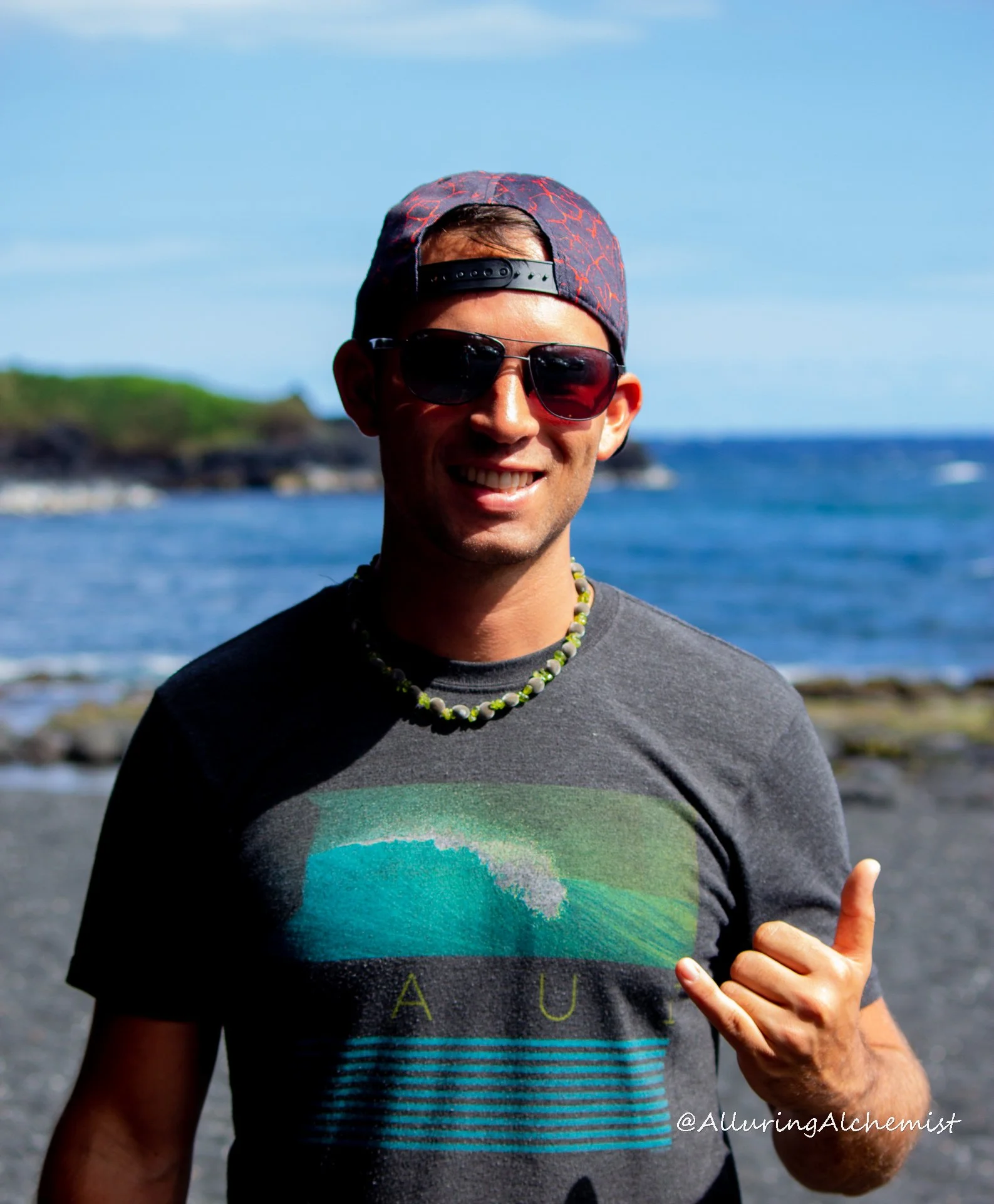 A man wearing sunglasses and a cap making a Shaka hand sign at the beach with water and sky in the background.