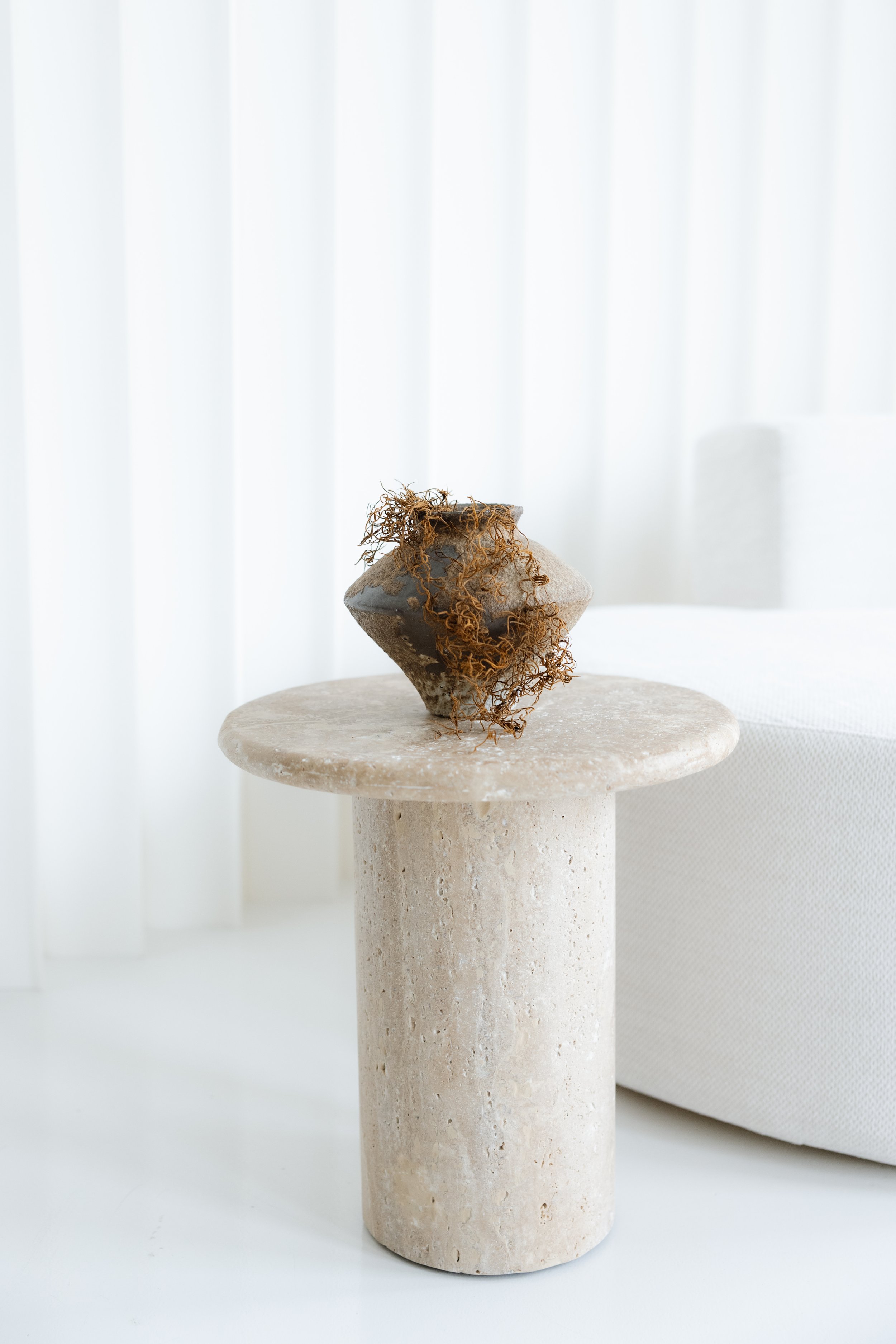 Minimalist beige stone side table with a textured, rounded top and cylindrical base, holding a geometric ceramic vase with dried, curly twigs, against a backdrop of white curtains and a white upholstered chair.