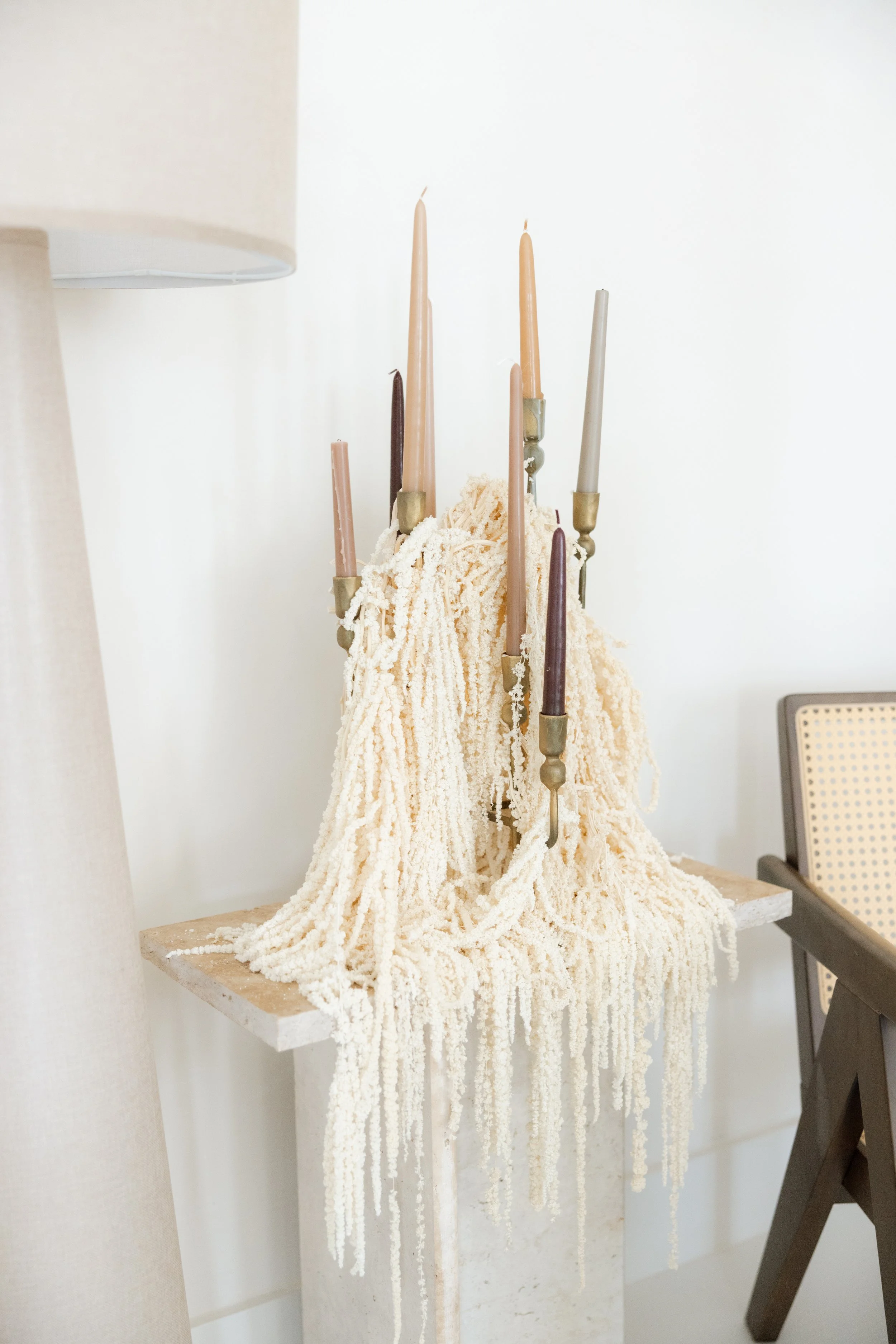 A candle chandelier with multiple pale pink, beige, and dark brown candles, draped with cream-colored textured yarn or fabric on a wooden side table, with a beige lamp and a beige and black chair nearby in a minimalistic room.