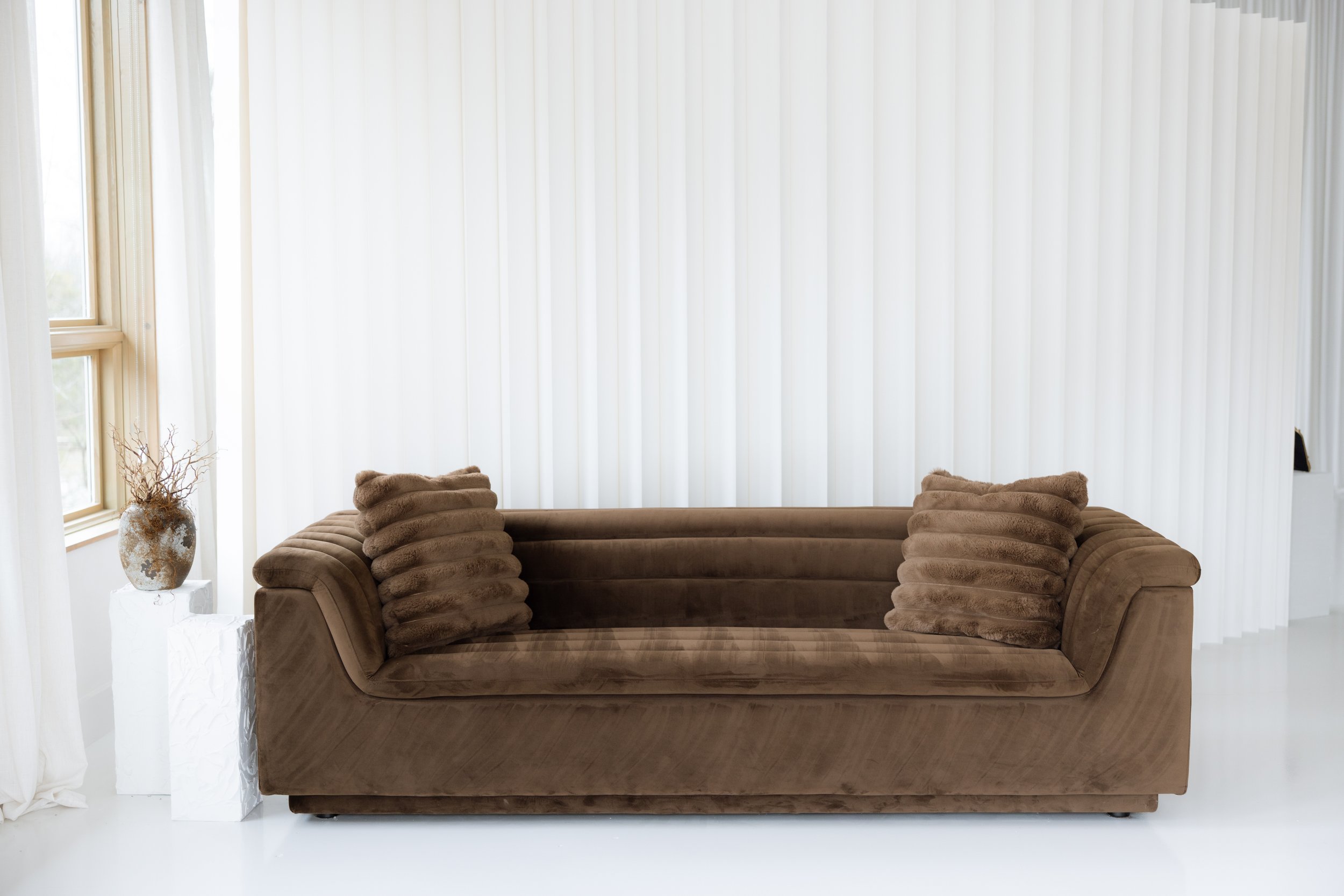A brown velvet sofa with two matching textured pillows in front of white vertical blinds and a window with cream-colored curtains, with a decorative vase on a white pedestal on the left side.