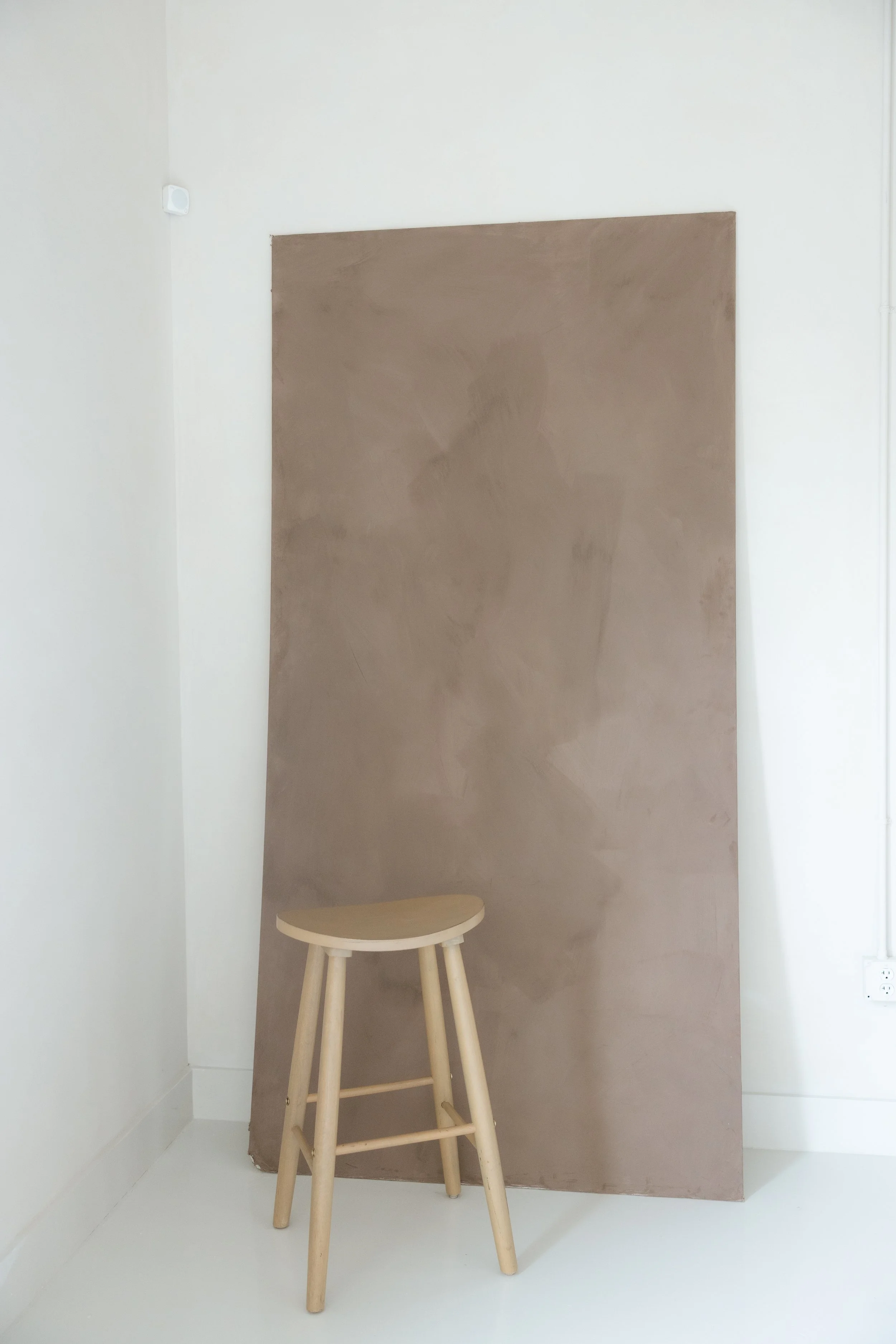 Wooden stool in front of a large, rectangular brown canvas on a white wall in a minimal room.