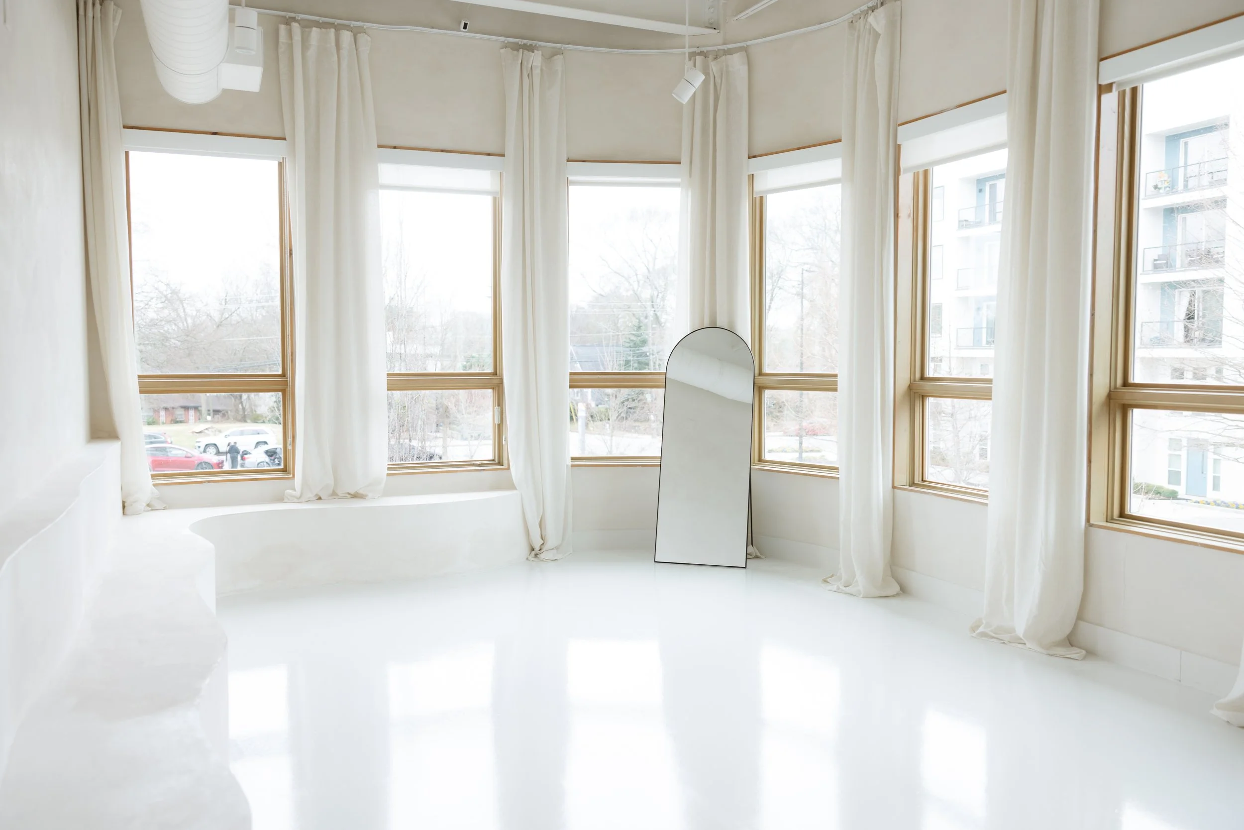Bright, minimalist room with large windows, white curtains, a full-length mirror, and white flooring.
