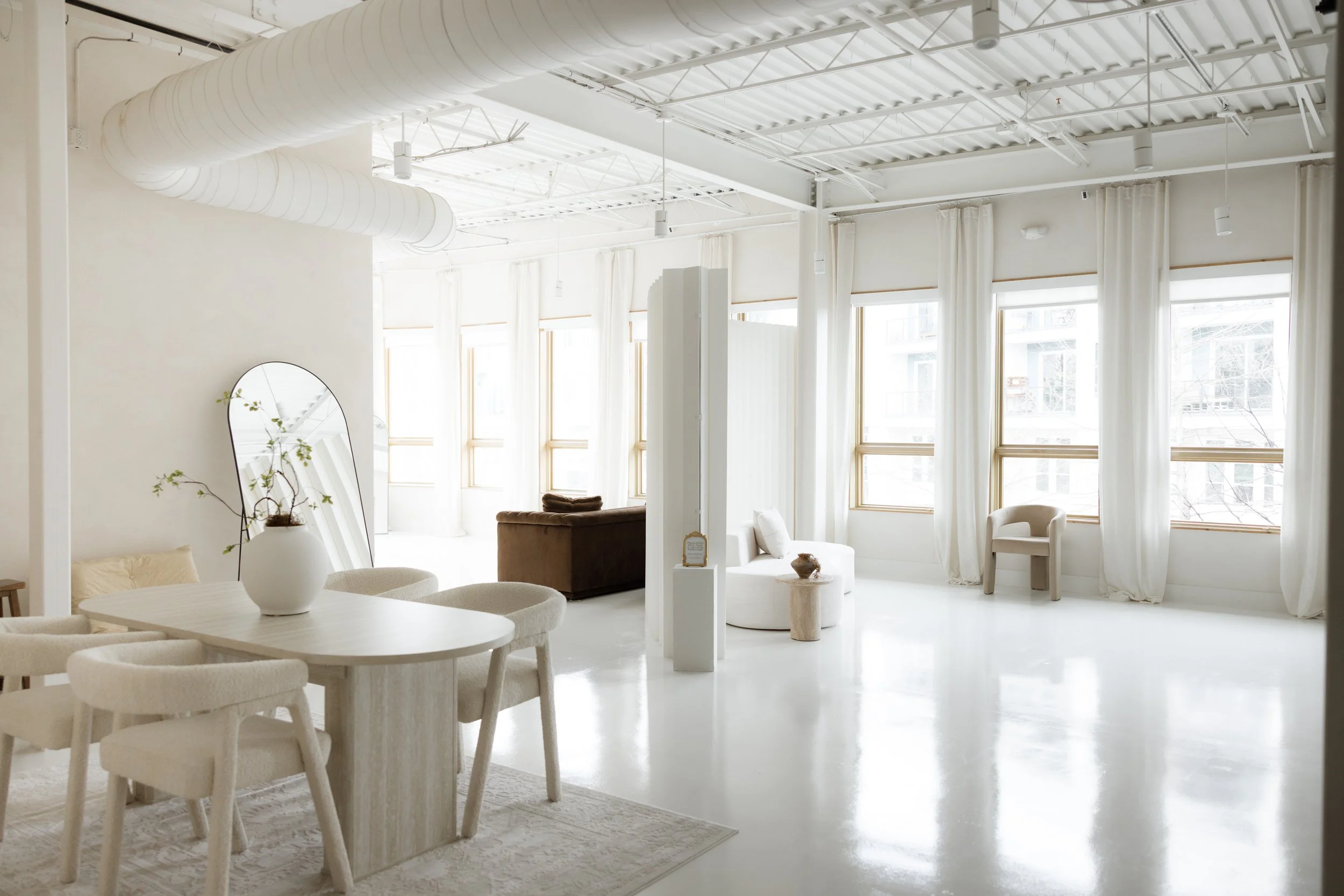 Bright, minimalist interior space with white walls, large windows with sheer curtains, white flooring, and simple furniture including a round table with chairs, a mirror, and various chairs and seating areas.