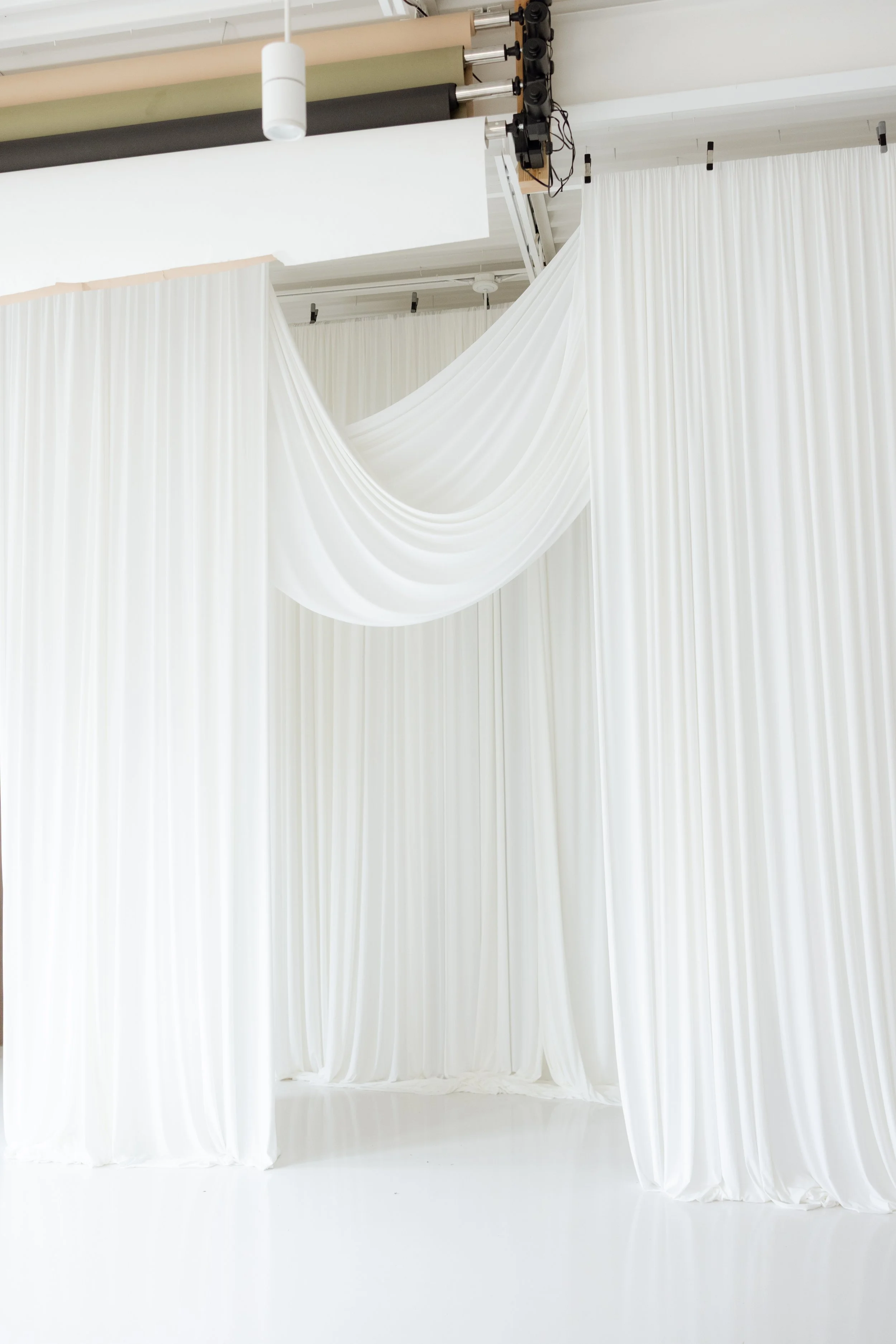White fabric draped as stage curtains with a white group of fabric panels hanging behind, set in a photo studio or event space with lighting equipment overhead.