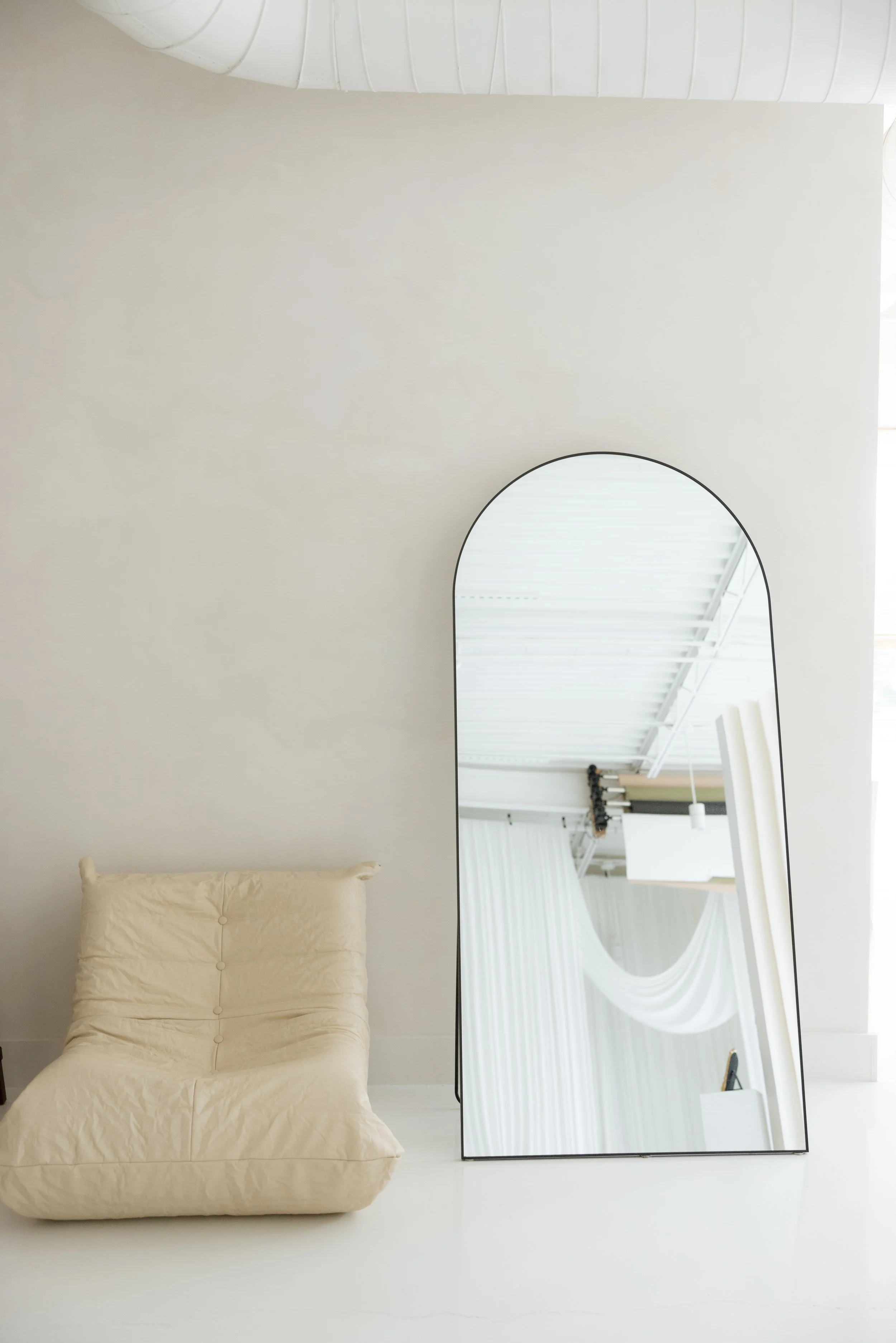 A beige floor chair with a tufted backrest next to a tall, arched mirror on a white floor against a plain white wall.