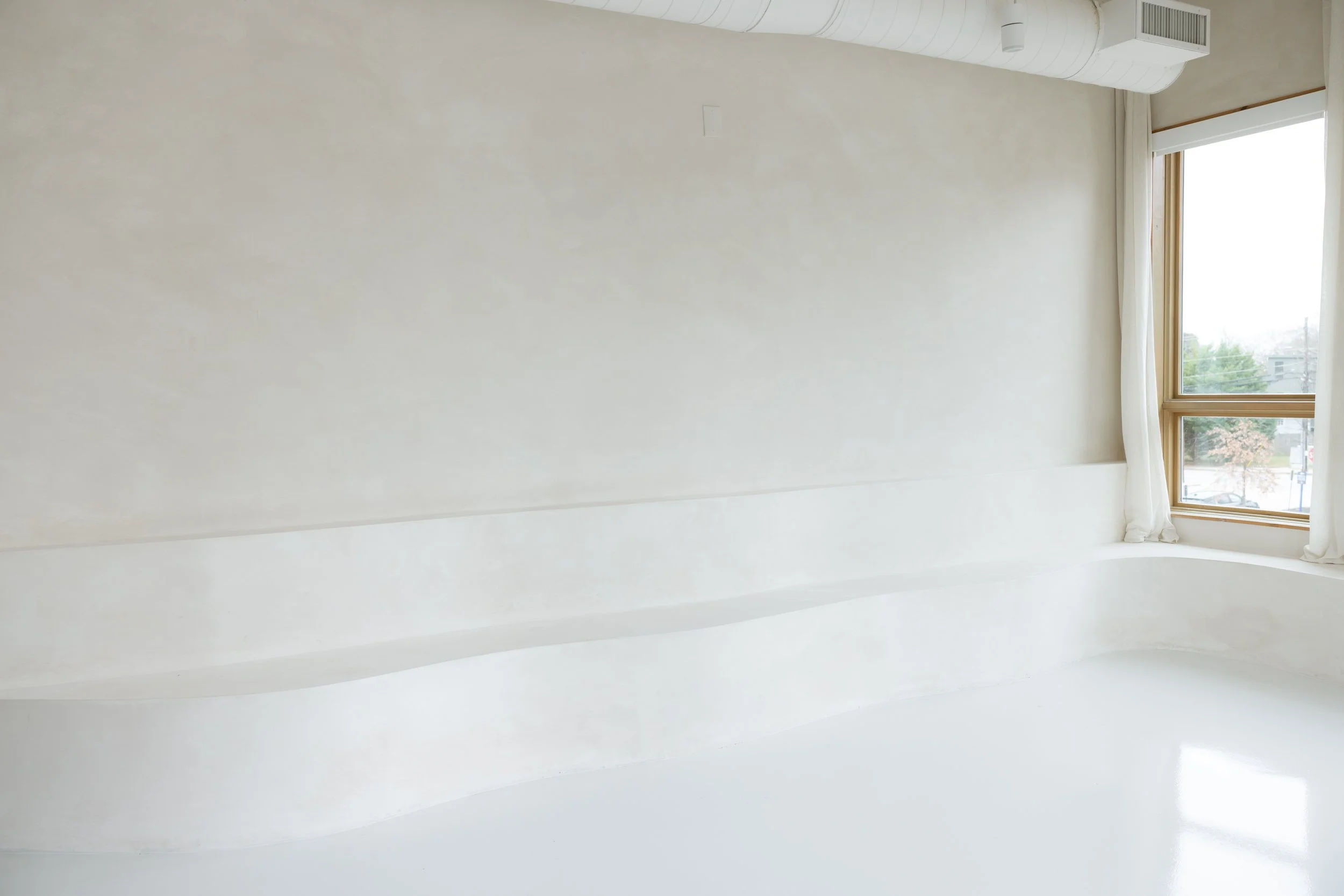 Empty room with white walls, large windows with curtains, and a curved built-in bench near the window.