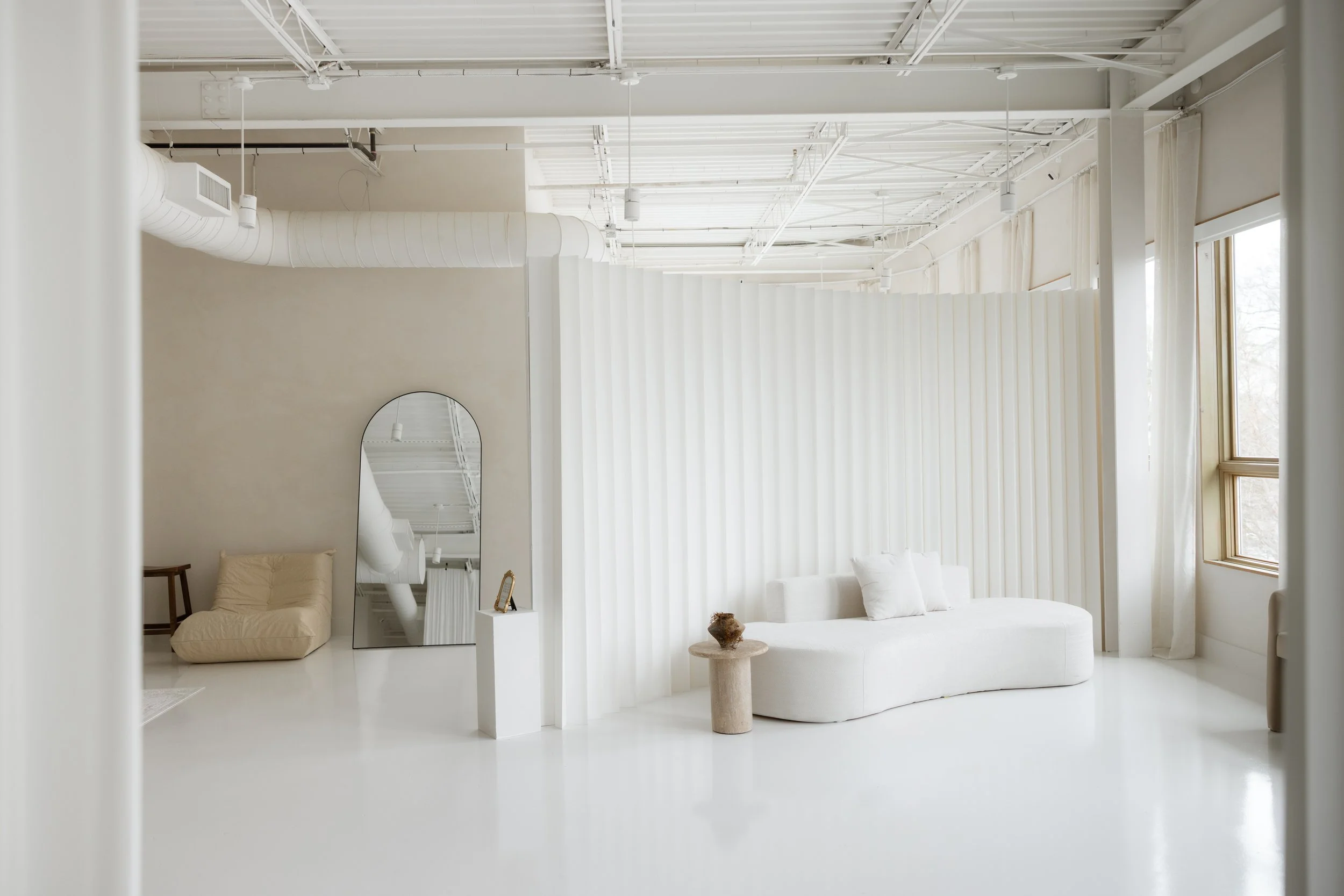 Minimalist indoor lounge with white walls, large windows, a curved white sofa, a small round side table with a decorative object, and a large standing mirror.