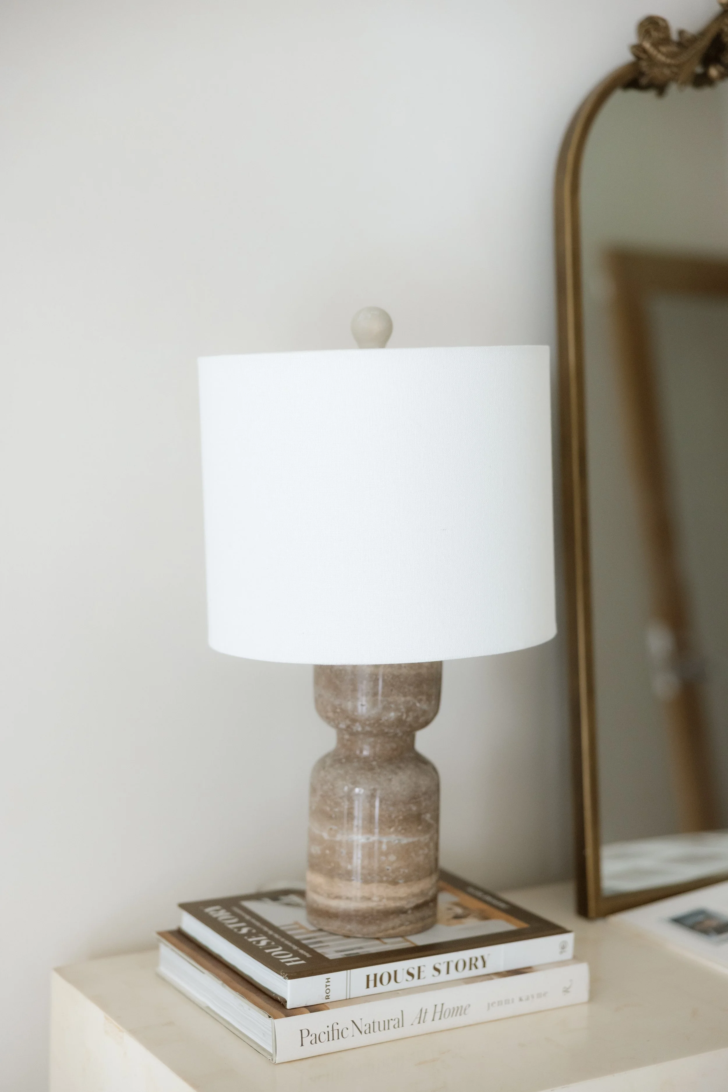 Table lamp with a beige textured base and a white lampshade, placed on two stacked books on a table next to a mirror.