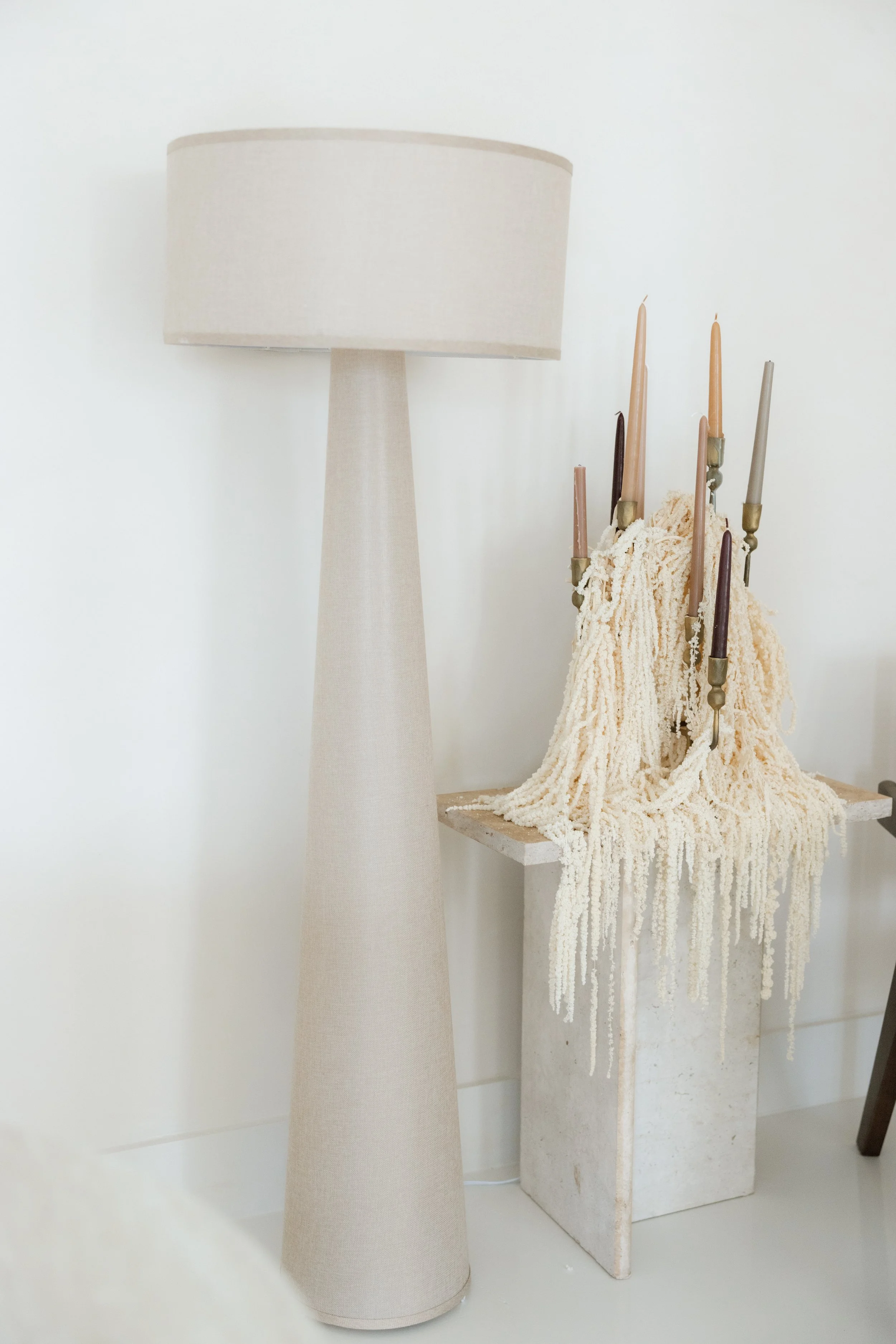 A cream-colored floor lamp with a tall, tapered base and a large cylindrical shade next to a small table holding dried flowers and candles.