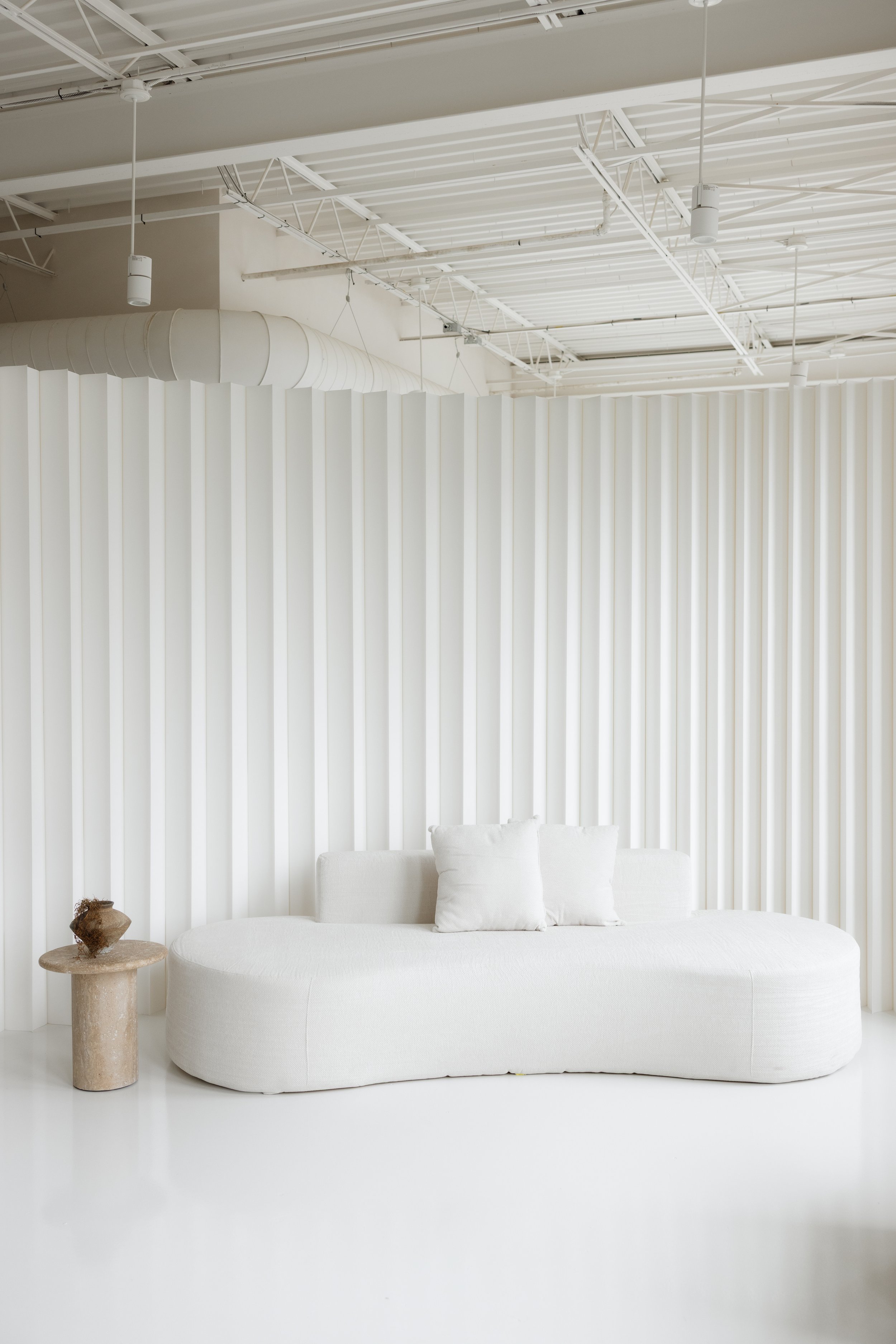 A modern, minimalist white sofa with two pillows in front of a white textured wall, beside a small round beige side table with a small decorative object, in a bright, spacious room with white ceiling and ductwork.