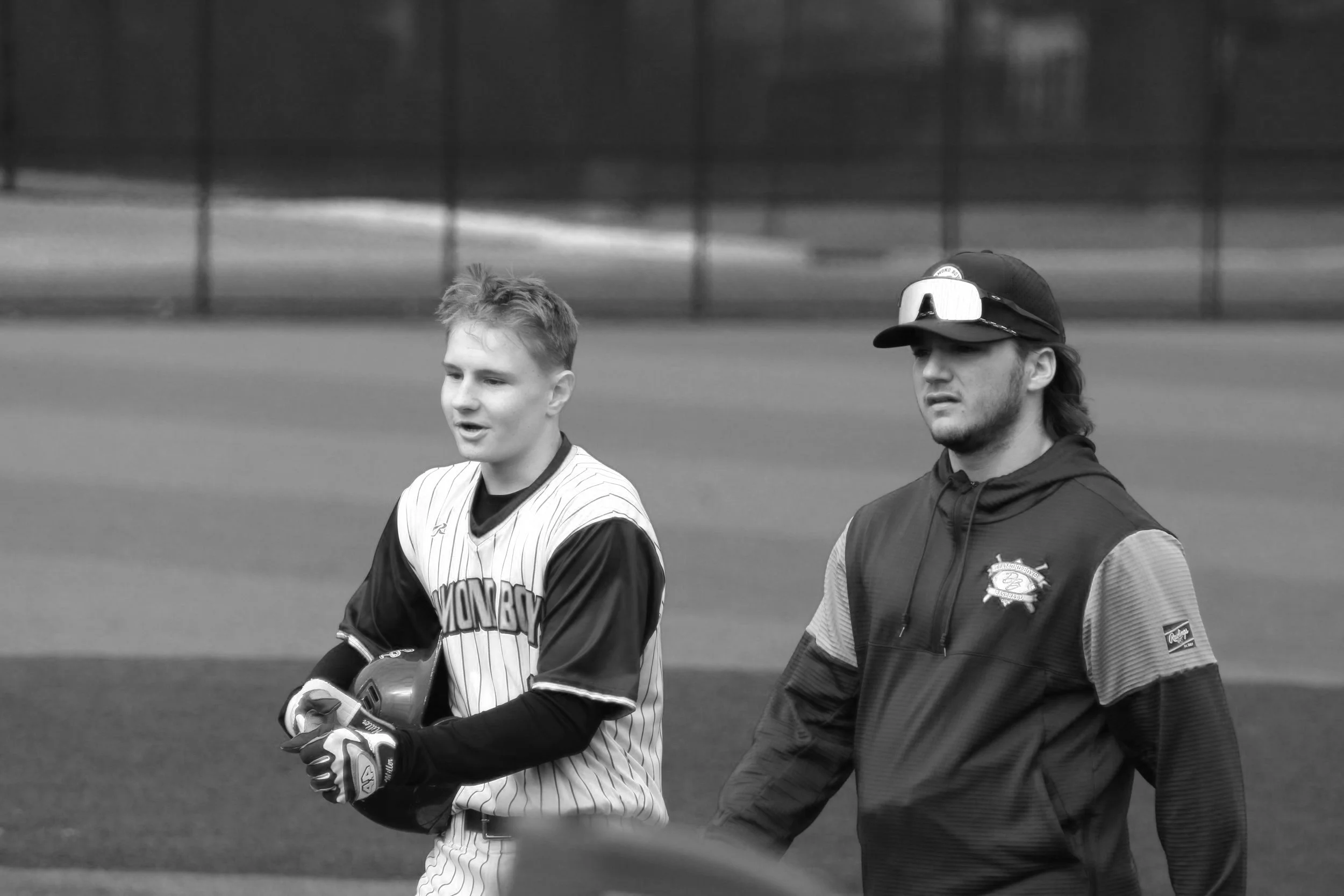 Two young men on a baseball field, one wearing a striped uniform holding a baseball glove, the other in a hoodie and cap, walking across the field.