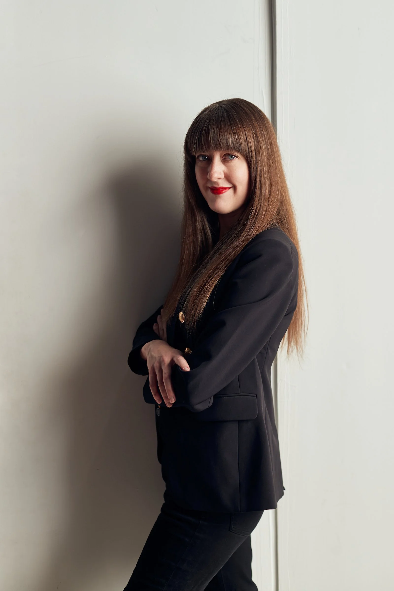 A woman with long brown hair and bangs, wearing a black blazer and pants, standing with her arms crossed and strong in stature against a plain wall.