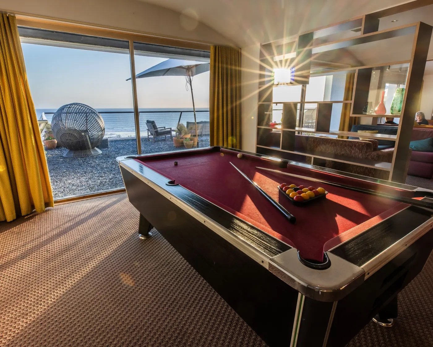 Indoor room with a red pool table near large glass sliding doors opening to an outdoor patio with chairs and an umbrella, overlooking the ocean at sunset.