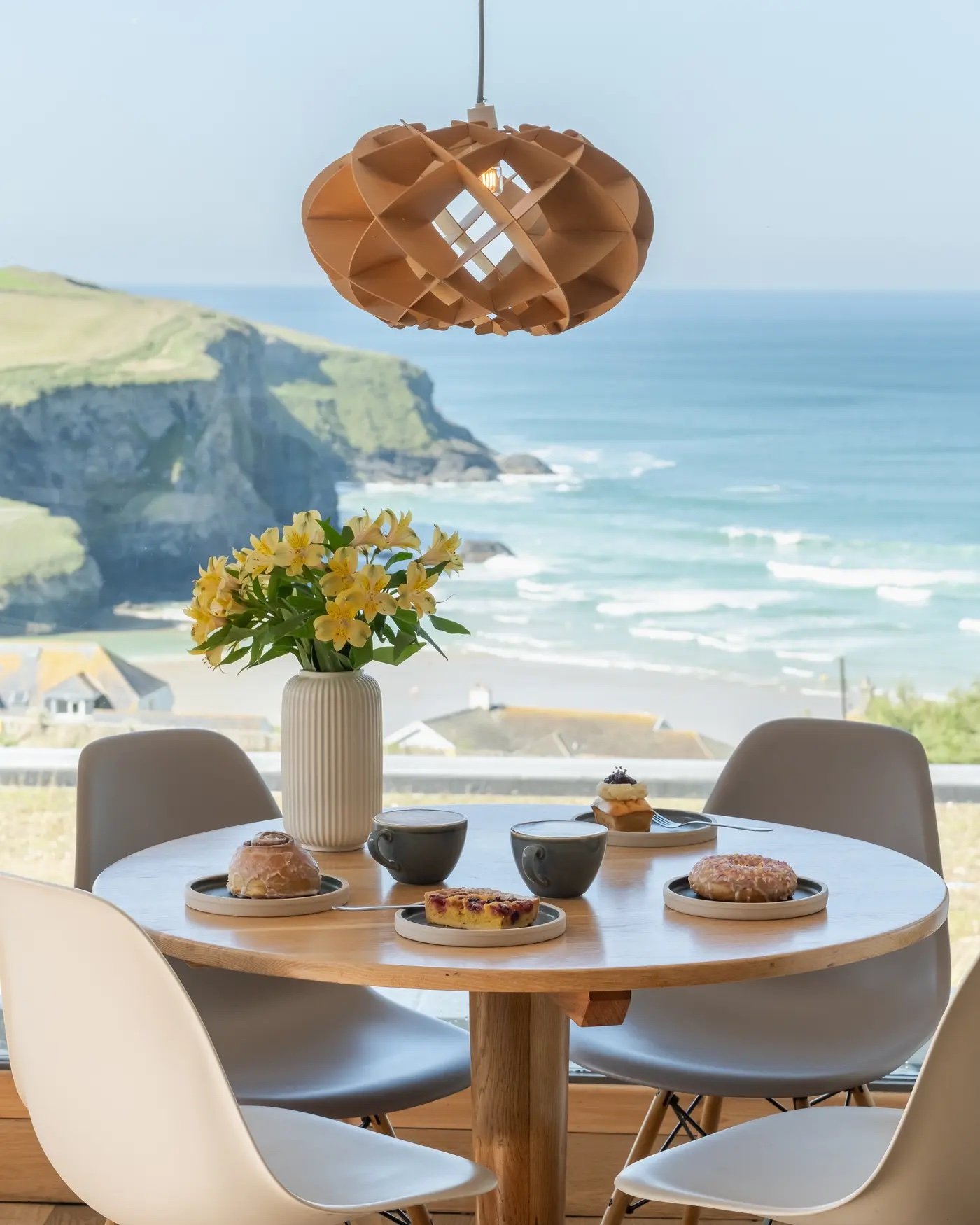 A round wooden table with four plates of pastries, a vase with yellow flowers, and two cups, set in front of a large window with a coastal view of cliffs, an ocean, and a partly cloudy sky. A modern hanging light fixture is above the table.