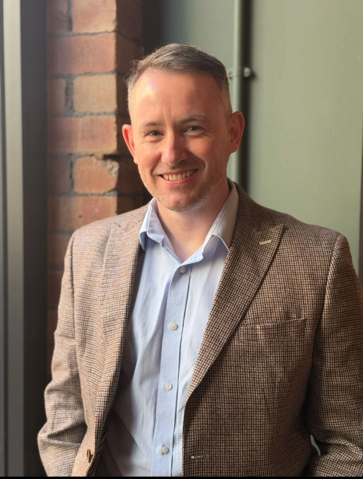 A man in a brown checkered blazer and light blue shirt smiling, standing indoors near a brick wall and green walls.
