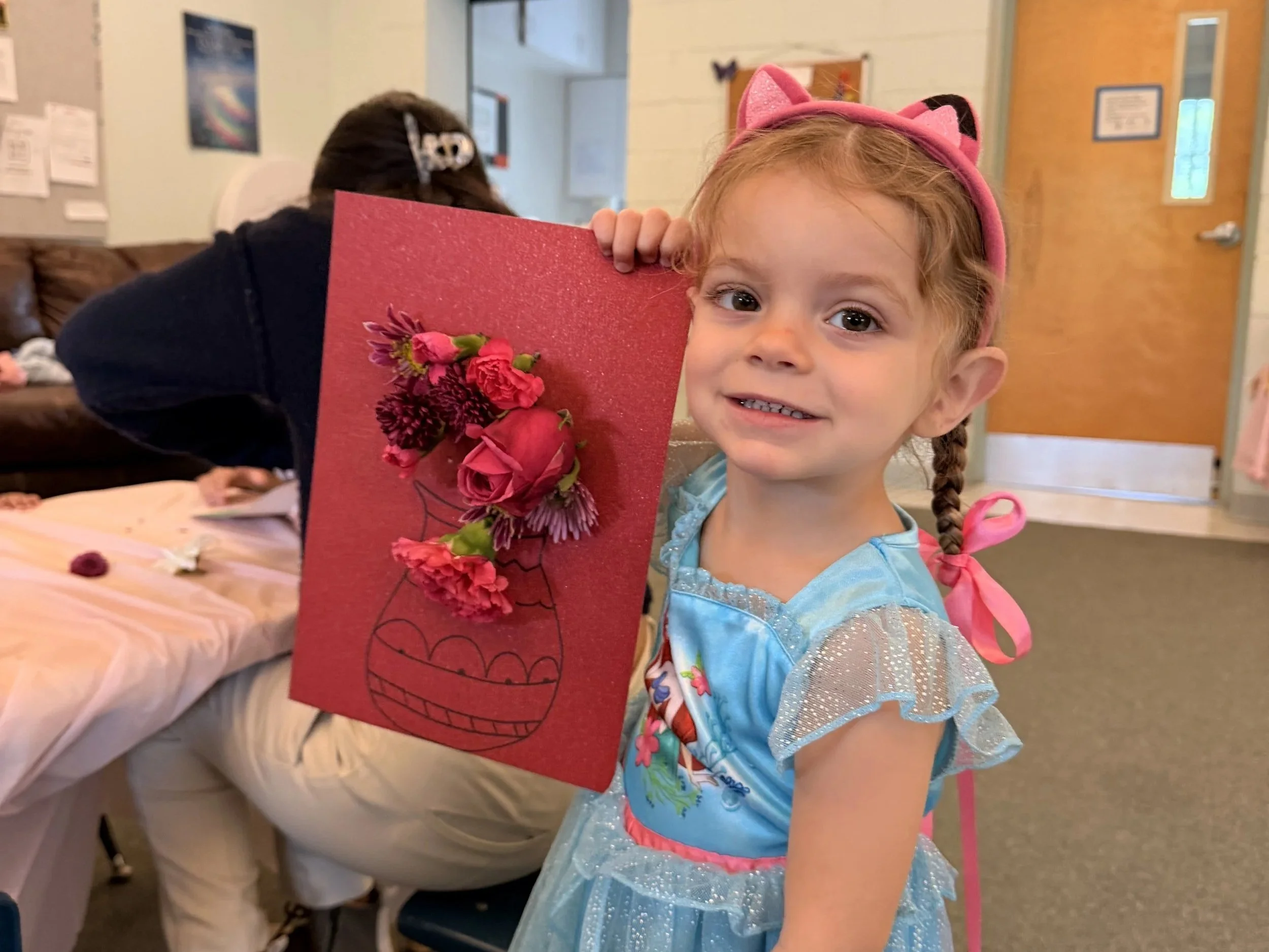 A young girl dressed as a princess holding a red card with pink and purple flowers on it, smiling at the camera in a room with wooden walls and a door.