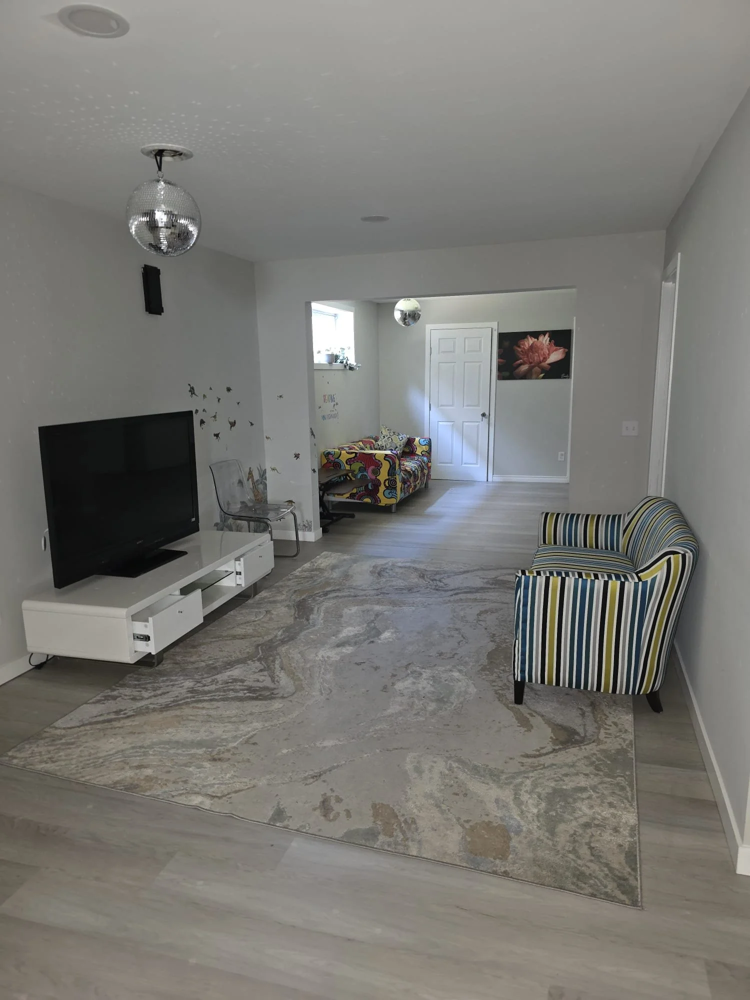 Living room with a large patterned area rug, striped armchair, wall-mounted TV, wall decals, and a colorful child's sofa near the door, with disco ball ceiling lights.