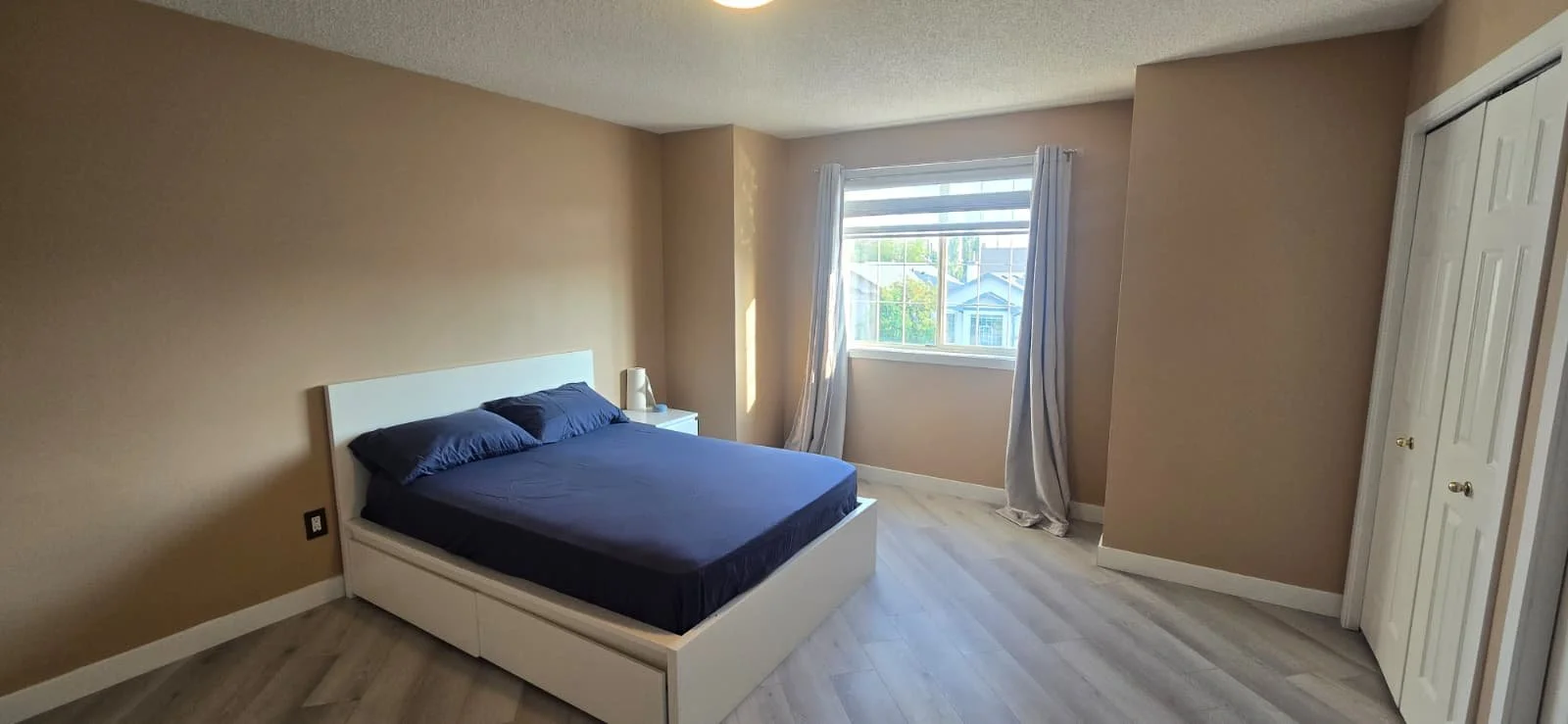 Empty bedroom with beige walls, white bed frame with dark bedding, two pillows, white nightstand, window with curtains allowing sunlight, closet with double doors.