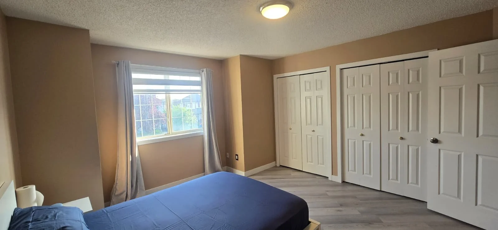 Bedroom with beige walls, a window with white curtains, a bed with blue bedding, and multiple white closets.