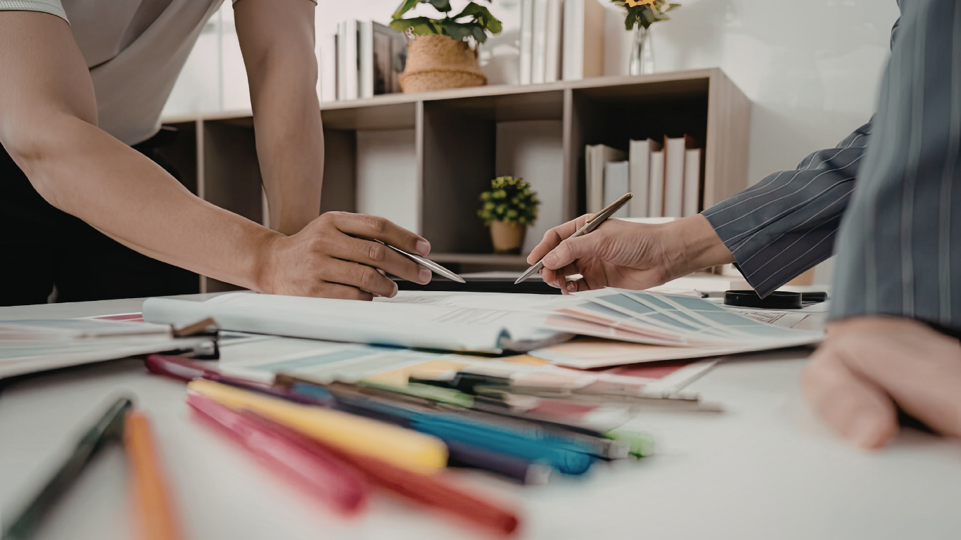Two people reviewing documents and color swatches at a desk with various colored markers and stationery.