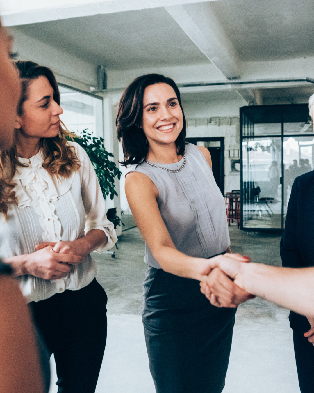 Woman smiling and shaking hands with another person in an office setting, with other women nearby.