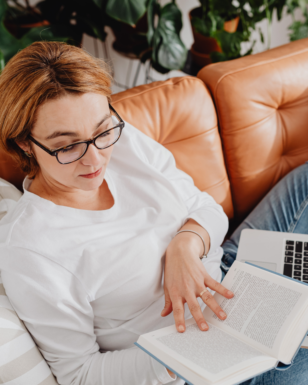 A woman with short red hair and glasses sitting on a tan leather couch, reading a book, with a laptop nearby and green plants in the background.