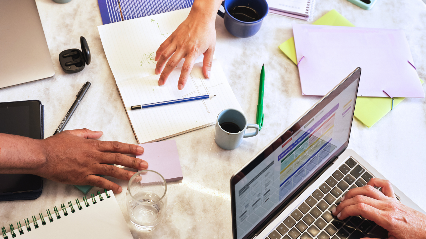 People working at a cluttered office desk with laptops, notebooks, pens, coffee cups, a glass of water, and various papers and folders.