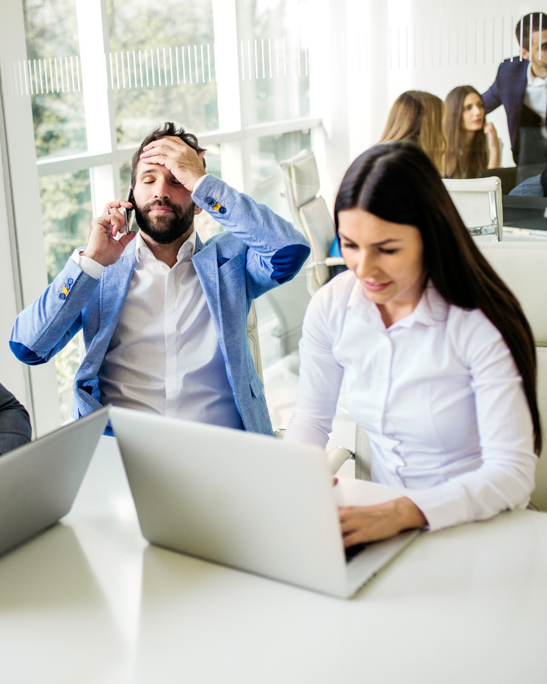 A frustrated man with a beard and a blue blazer sitting at a table, holding his forehead with one hand and talking on a phone with the other, while a woman in a white shirt works on a laptop.