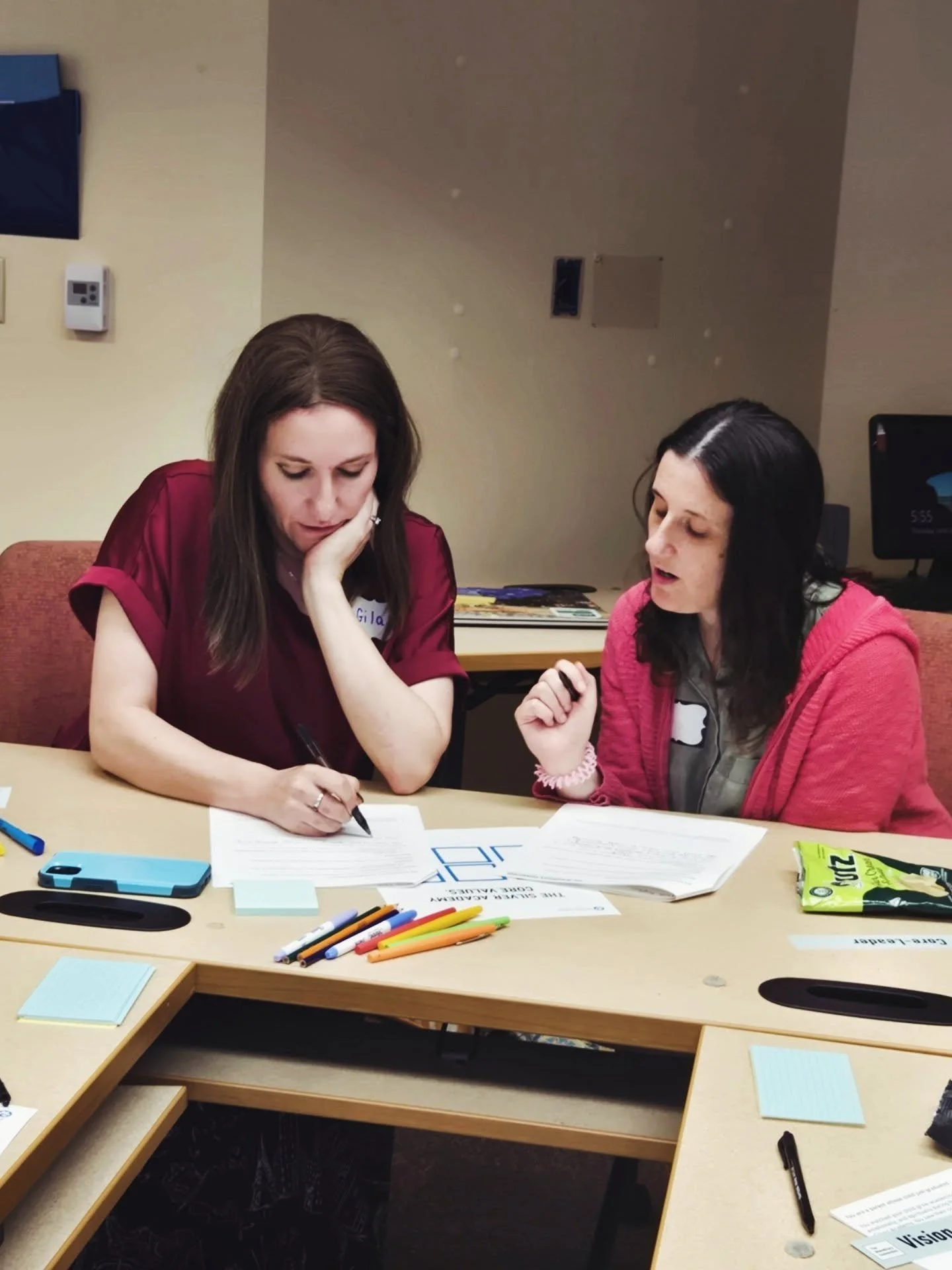 Two women sitting at a table looking at documents, surrounded by pens, sticky notes, and snacks, engaged in discussion.