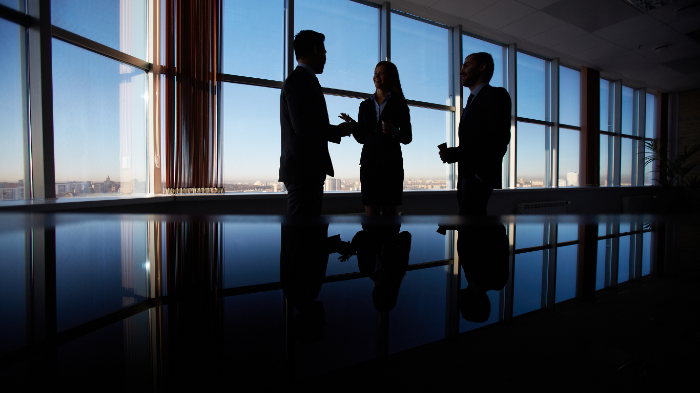 Three silhouetted business professionals in suits talking and sharing a drink in a modern office with large windows showing a city skyline.