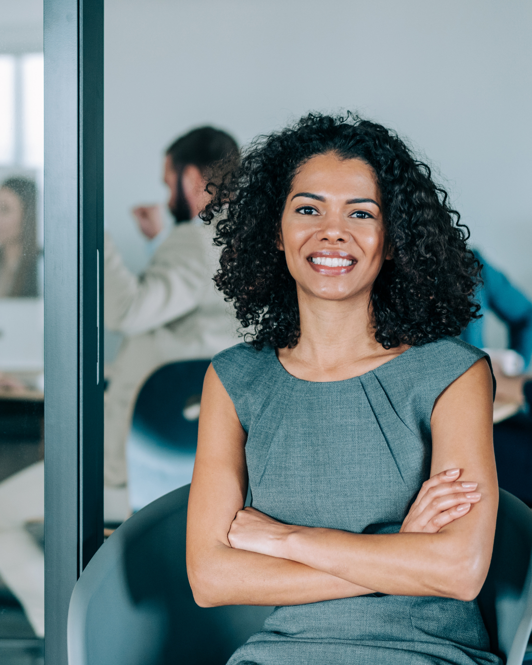 Professional woman with curly hair smiling and sitting with arms crossed in a modern office.