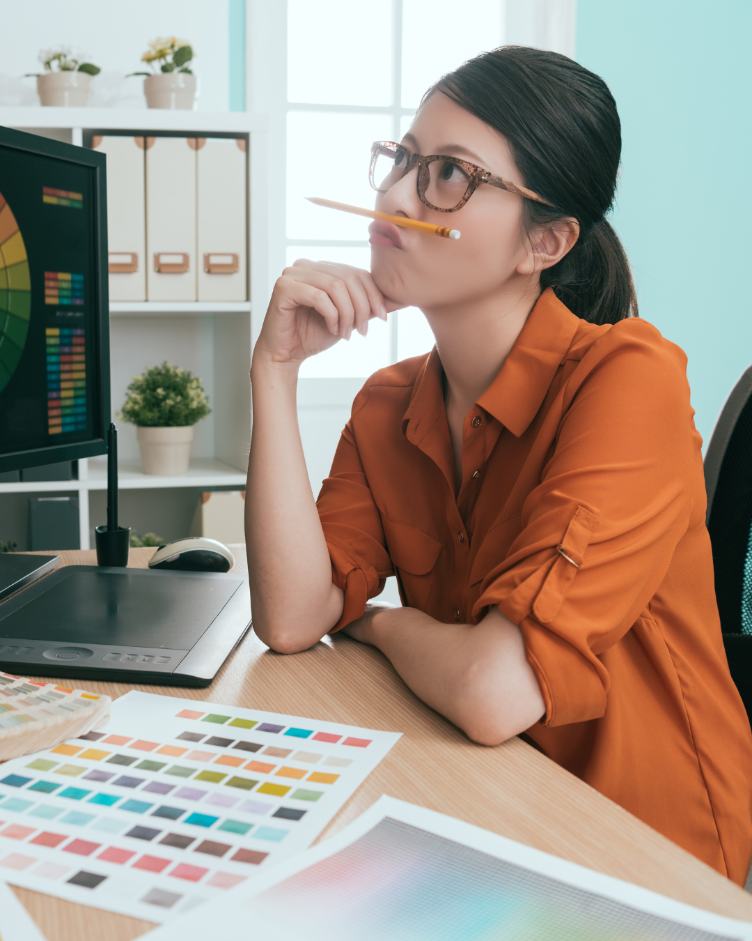 A woman with glasses and a pencil in her mouth, sitting at a desk with color swatches and a graphic tablet, thoughtfully working on a project in an office.