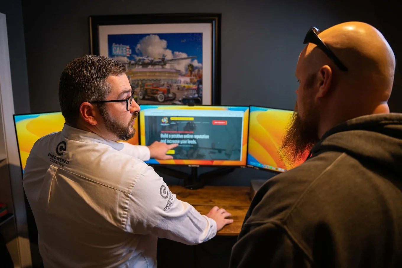 Two men sit at a desk in front of three computer monitors. One man with glasses and a beard points at his screen while the other man, with a bald head and beard, looks on. A framed picture of an airplane and cars is on the wall behind them.
